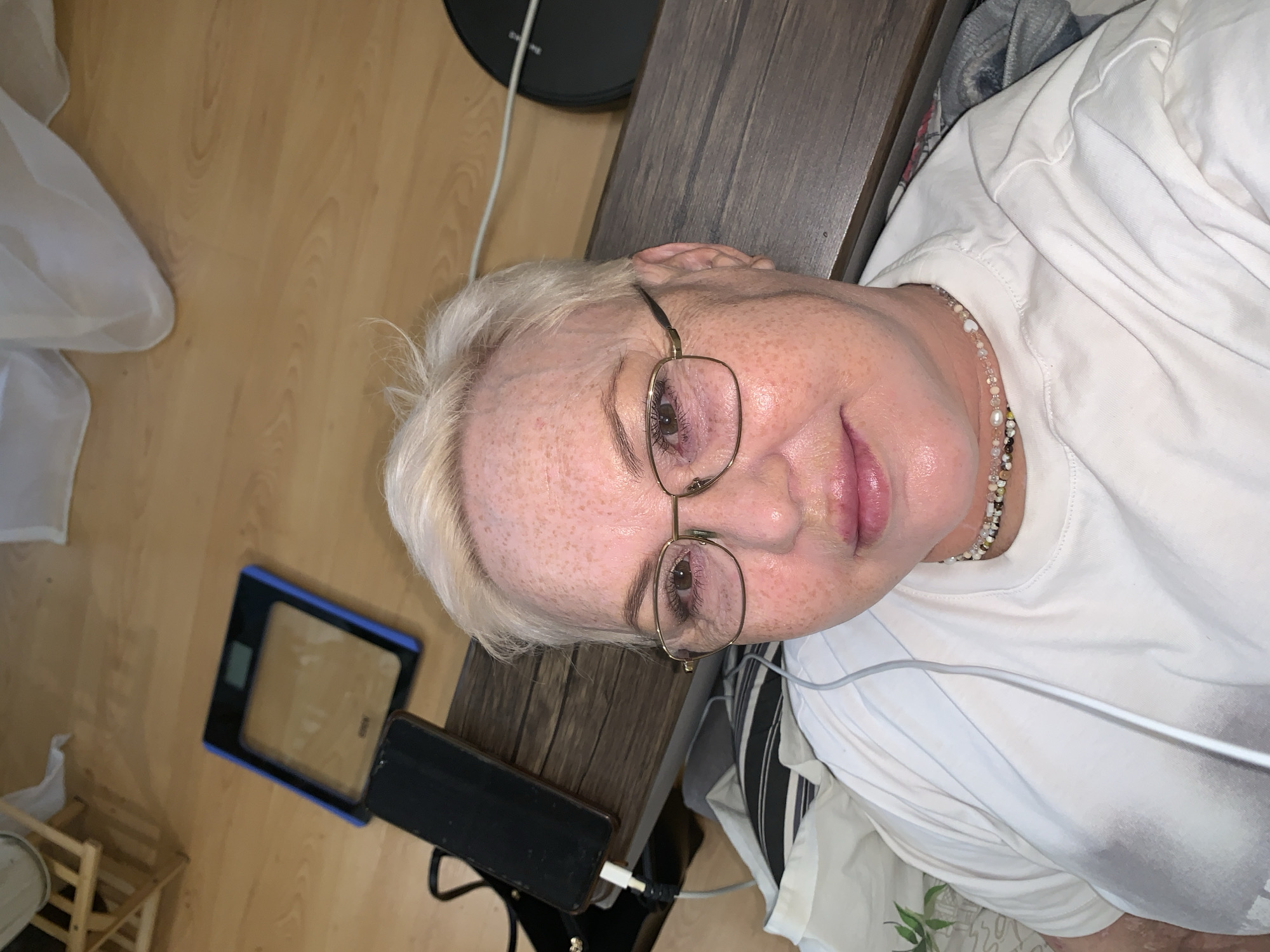 A middle-aged Caucasian woman with freckled skin and short, platinum blonde hair is lying on her side, wearing glasses, a white t-shirt, and a beaded necklace. Her head rests on a dark wooden surface resembling a bedside table. Next to the table, on a wooden floor, are a digital scale, a black mobile phone connected to a charging cable, and a robotic vacuum cleaner. A gauzy white curtain is partially visible in the lower left corner. The location in the city of Chaikovsky, Russia cannot be confirmed based solely on this image.