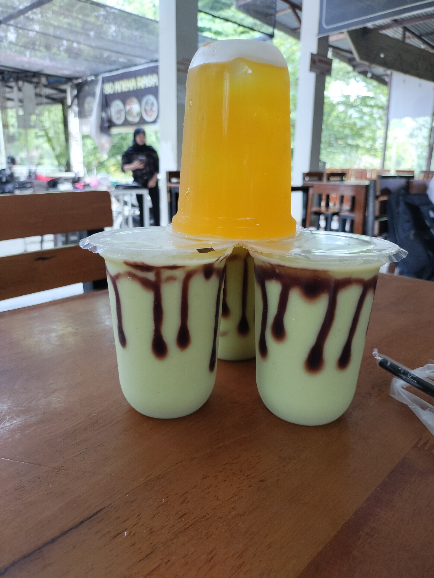 Four plastic cups of beverages are arranged on a wooden table. Three cups are in the foreground, each filled with a light green liquid and decorated with dark brown drizzles on the inside of the cup. These three cups are topped with clear plastic lids. A fourth cup, filled with a bright yellow liquid and also topped with a clear plastic lid, is stacked on top of the three foreground cups. The background appears to be an outdoor seating area, possibly a cafe or restaurant, with tables, chairs, and greenery visible. A person wearing dark clothing and a headscarf is standing in the middle ground, looking at a phone. A banner with text and images is hanging in the background. The scene is set in Keudealue Ie Puteh, Indonesia.