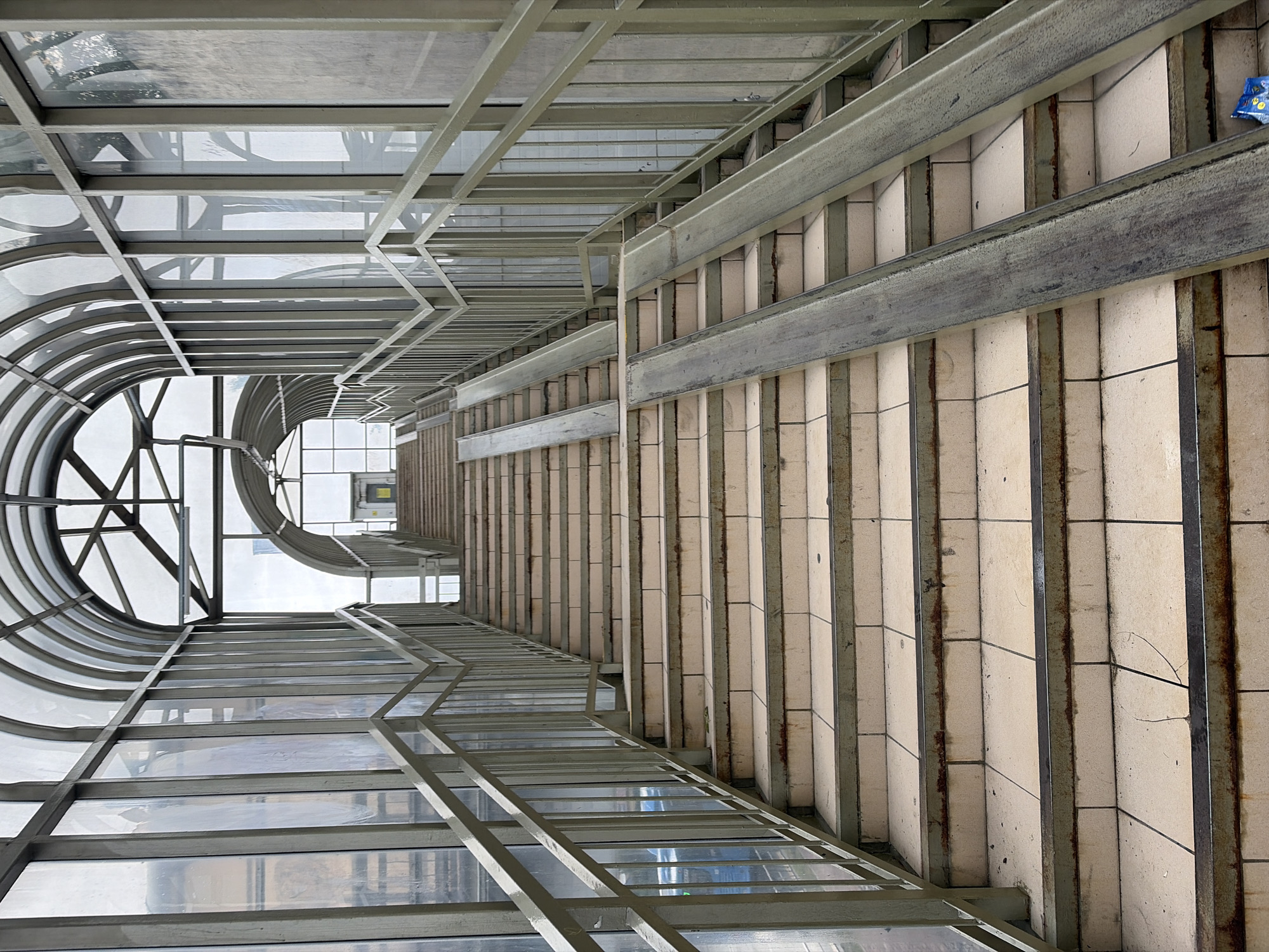The image depicts an elevated walkway or overpass in Sochi, Russia. The walkway is enclosed with a metal frame and glass or transparent panels. The structure forms a curved tunnel at one end, providing a view of the sky. On the right, there is a staircase with light-colored, rectangular tiles and metal railings. A blue wrapper or object is visible in the top right corner of the image. The overall impression is an industrial aesthetic, with a focus on the structural elements of the walkway.