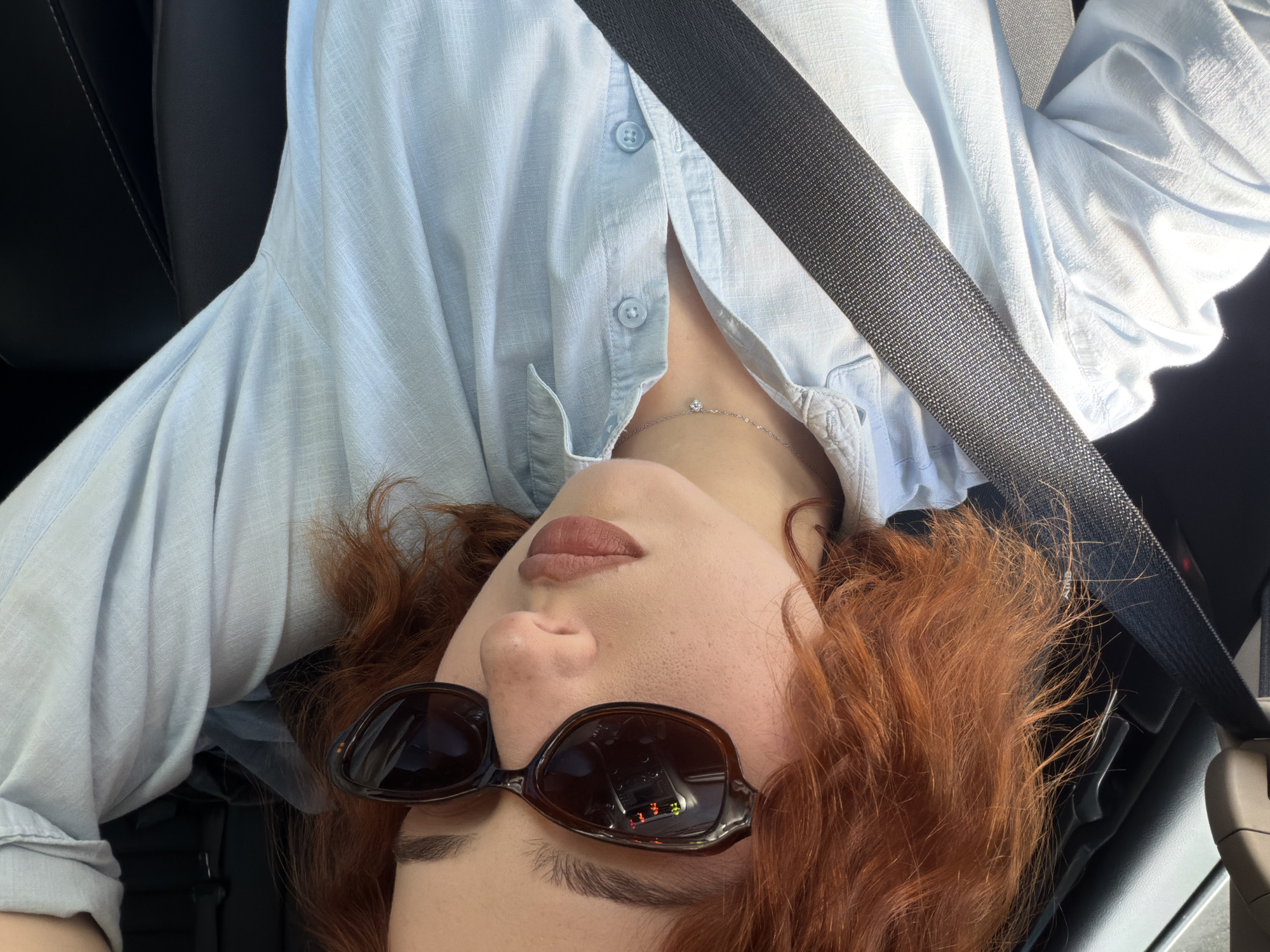The image shows a person with red hair wearing sunglasses and a light blue button-down shirt. They are wearing a seatbelt, suggesting they are in a car. A silver necklace is visible. The reflection in their sunglasses appears to show a car dashboard. The location, Tbilisi, Georgia, cannot be determined from the information in the image.