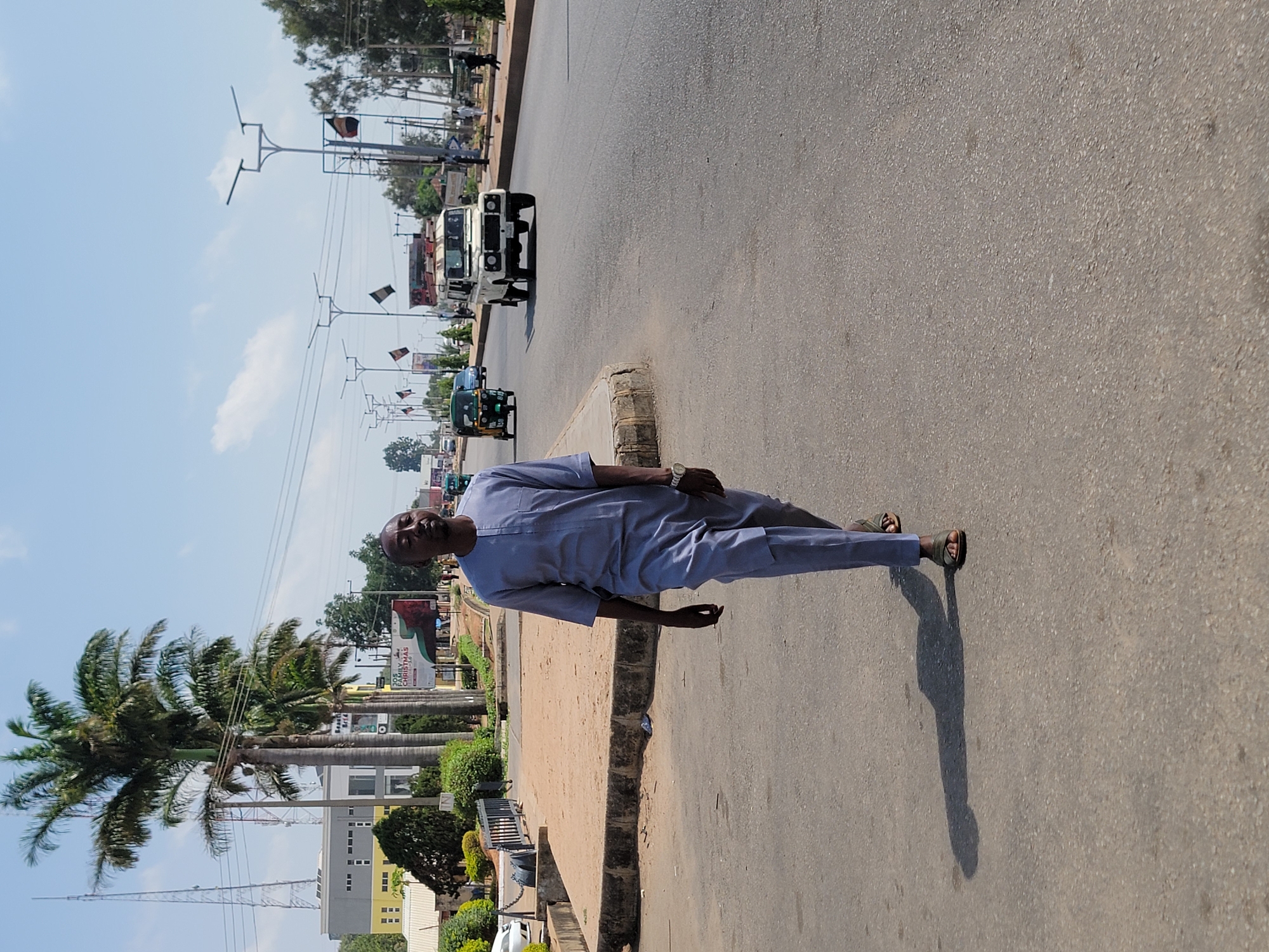 An adult Black male stands on a paved road in Jos, Nigeria. He wears a light blue traditional tunic and pants. He is wearing sandals. The surrounding environment includes tall palm trees, a building with windows, power lines, and various vehicles on the road. The sky is blue with some clouds. There are street lights and flags visible. A white pickup truck is driving on the road.
