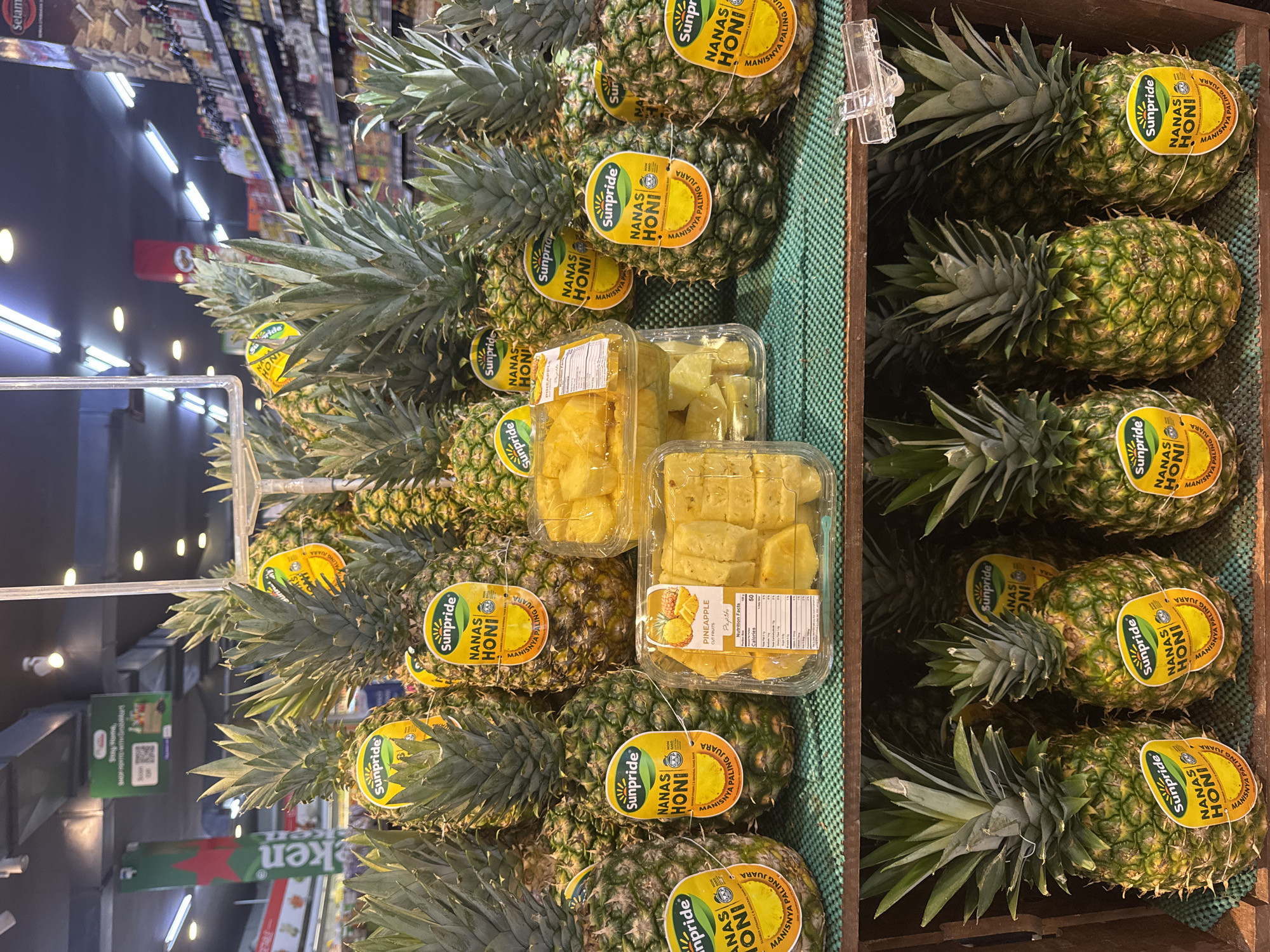 The image depicts a produce display in what appears to be a supermarket in Kuta Selatan, Indonesia. The main subjects are a large quantity of fresh pineapples, both whole and pre-cut, arranged on shelves and in wooden crates. Many of the whole pineapples have yellow circular stickers with the brand "Sunpride" and text in Indonesian, indicating they are "Nanas Honi" and "Manisnya Paling Juara."
In the foreground, several pineapples are visible in a wooden crate, with their spiky green crowns facing upwards. To the right of these, there are plastic containers filled with cut pineapple pieces. Behind this, on a higher shelf, more whole pineapples are displayed, also with the Sunpride stickers. In the background, shelves stocked with various products are visible, along with ceiling lights and signage. One sign in the upper left corner is green with a QR code and text, suggesting a promotional or informational display. The overall environment suggests a retail setting focused on fresh produce. There are no people visible in the image.