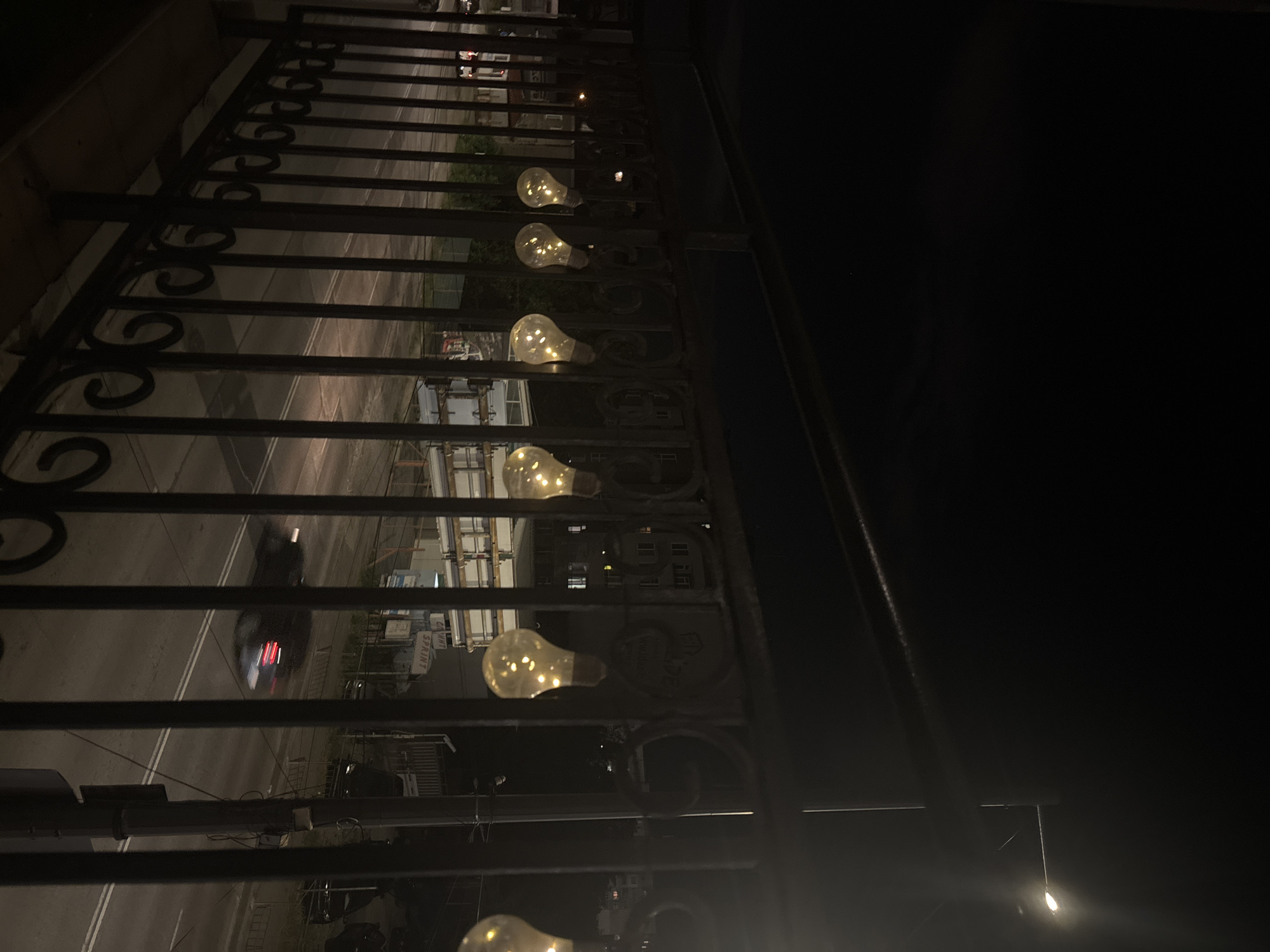 This image presents a detailed view of an urban night scene captured from an elevated position, likely an overpass or bridge. In the immediate foreground, a dark, ornate metal railing with vertical bars and decorative scrollwork at its base spans the width of the frame. Six illuminated decorative lightbulbs, emitting a warm, diffused glow from internal filaments, are suspended along the upper portion of this railing.
Below the railing, a multi-lane asphalt road stretches into the distance, delineated by clear white double lines. A dark sedan is visible in motion on the road, exhibiting significant motion blur, with its taillights brightly illuminated. Further along the street to the left, a white vehicle is partially discernible, possibly parked.
Along the left side of the street, several buildings are present. A prominent white sign clearly displays "САНИТ" (SANIT) above "СПРИНТ" (SPRINT) in Cyrillic script. Below these, a smaller sign reads "РЕДИ-М" (REDI-M). Further into the midground, multi-story residential buildings with some illuminated windows are visible. On the right side of the street, a tall street light post with a single, intensely bright lamp casts a strong beam of light, creating lens flare effects. Various other commercial structures and what appear to be construction scaffolding are visible beyond the street. The background consists of a dark, clear night sky.
The presence of Cyrillic script on the signs, specifically "САНИТ", "СПРИНТ", and "РЕДИ-М", unequivocally places this scene in a Bulgarian-speaking location. The urban architecture, infrastructure, and general environment are consistent with typical street views found within the city of Sofia, Bulgaria. No discernible individuals are present in the frame. The overall atmosphere is that of a bustling city at night, illuminated by various artificial light sources.