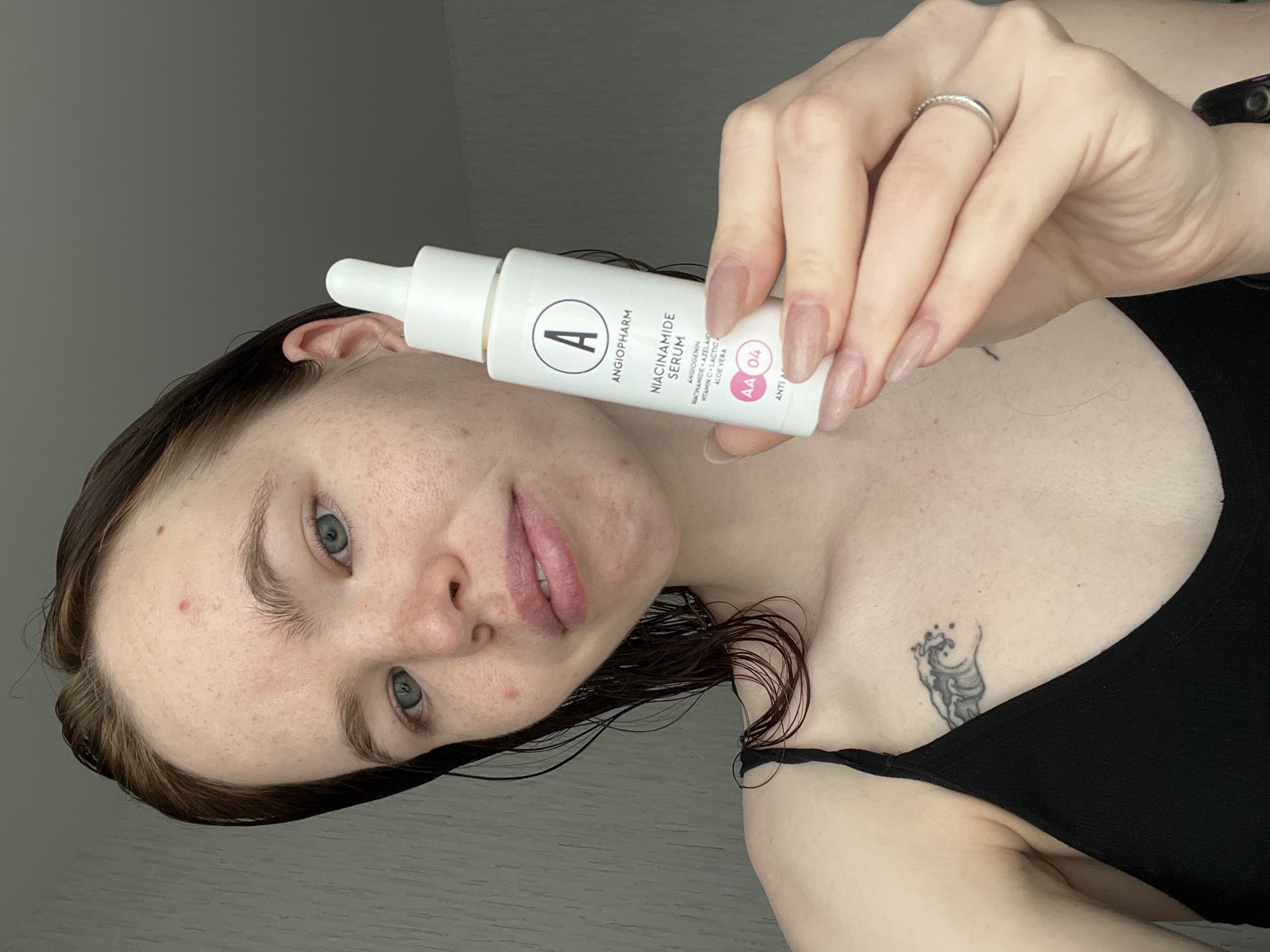A woman is holding a white bottle of Angiopharm Niacinamide Serum close to her face. She is wearing a black tank top, and a small tattoo is visible on her chest. She is also wearing a ring on her finger. Her hair is wet, and she has some skin imperfections. The background is a light gray wall. There is no indication the photo was taken in Perm, Russia.