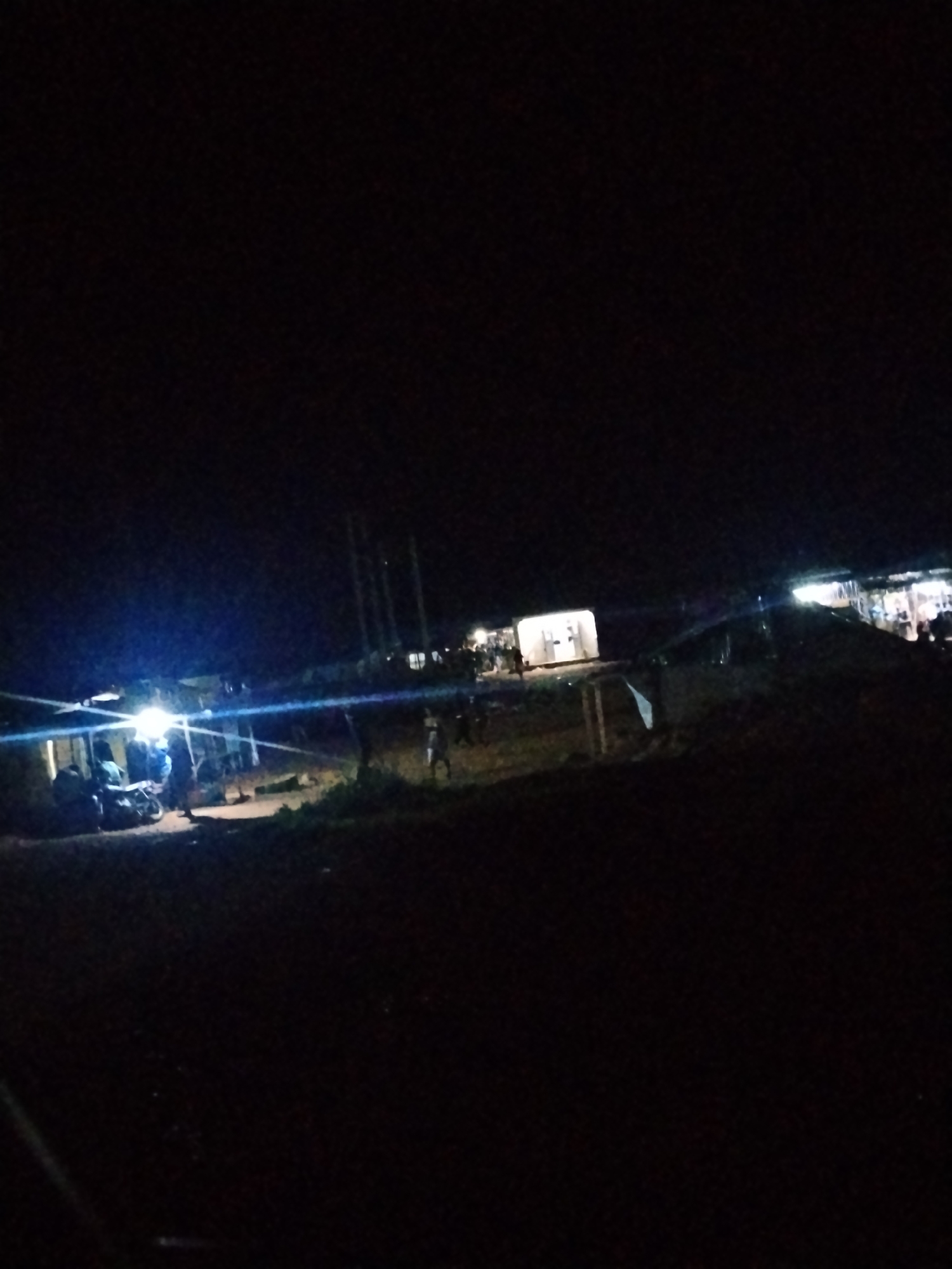 The image depicts a nighttime scene in Durumi, Nigeria, likely a market or gathering place, illuminated by artificial lights. In the foreground on the left, a motorcycle is visible, with several individuals gathered around it, suggesting a potential interaction or pause. Further in the background, structures resembling stalls or shops are present, with lights indicating activity within them. The overall environment appears to be an outdoor setting, with unpaved ground visible. Silhouettes of people can be seen moving in and around these structures. The lighting creates harsh shadows and highlights, typical of a nighttime environment with limited illumination sources. The scene appears somewhat chaotic due to the low light conditions and the presence of multiple individuals and structures.