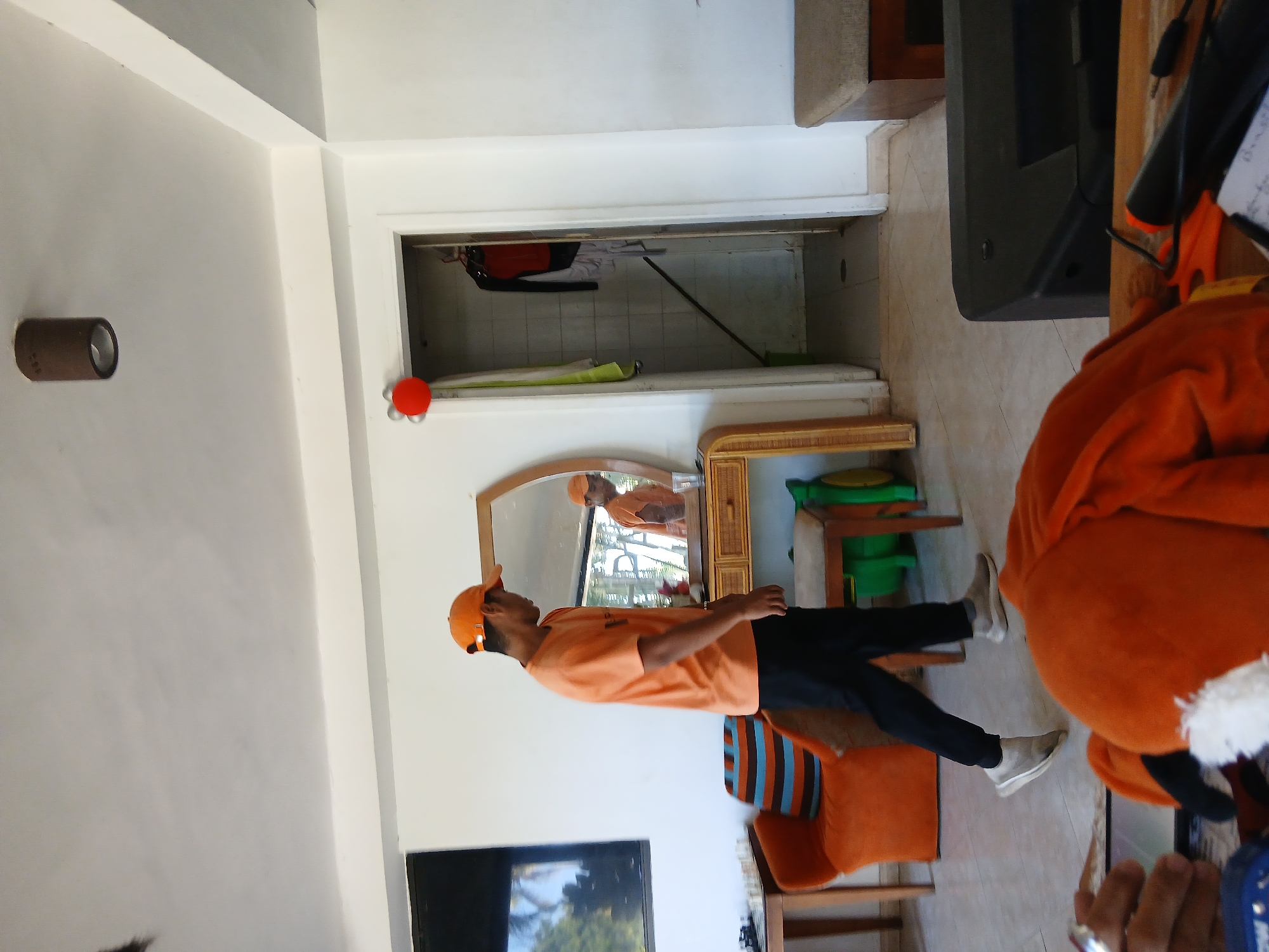 A person wearing an orange cap and shirt stands in a room, facing a mirror. The person's reflection is visible in the mirror. The room has white walls and a tiled floor. There is a dresser with a wicker front and a mirror on top. Behind the dresser, there is a doorway leading to another room. In the foreground, part of an orange stuffed animal is visible. The person appears to be looking at their reflection in the mirror. The time of day is unclear, but the lighting suggests it is daytime. The setting appears to be a house or an indoor space. There is no visible text in the image.