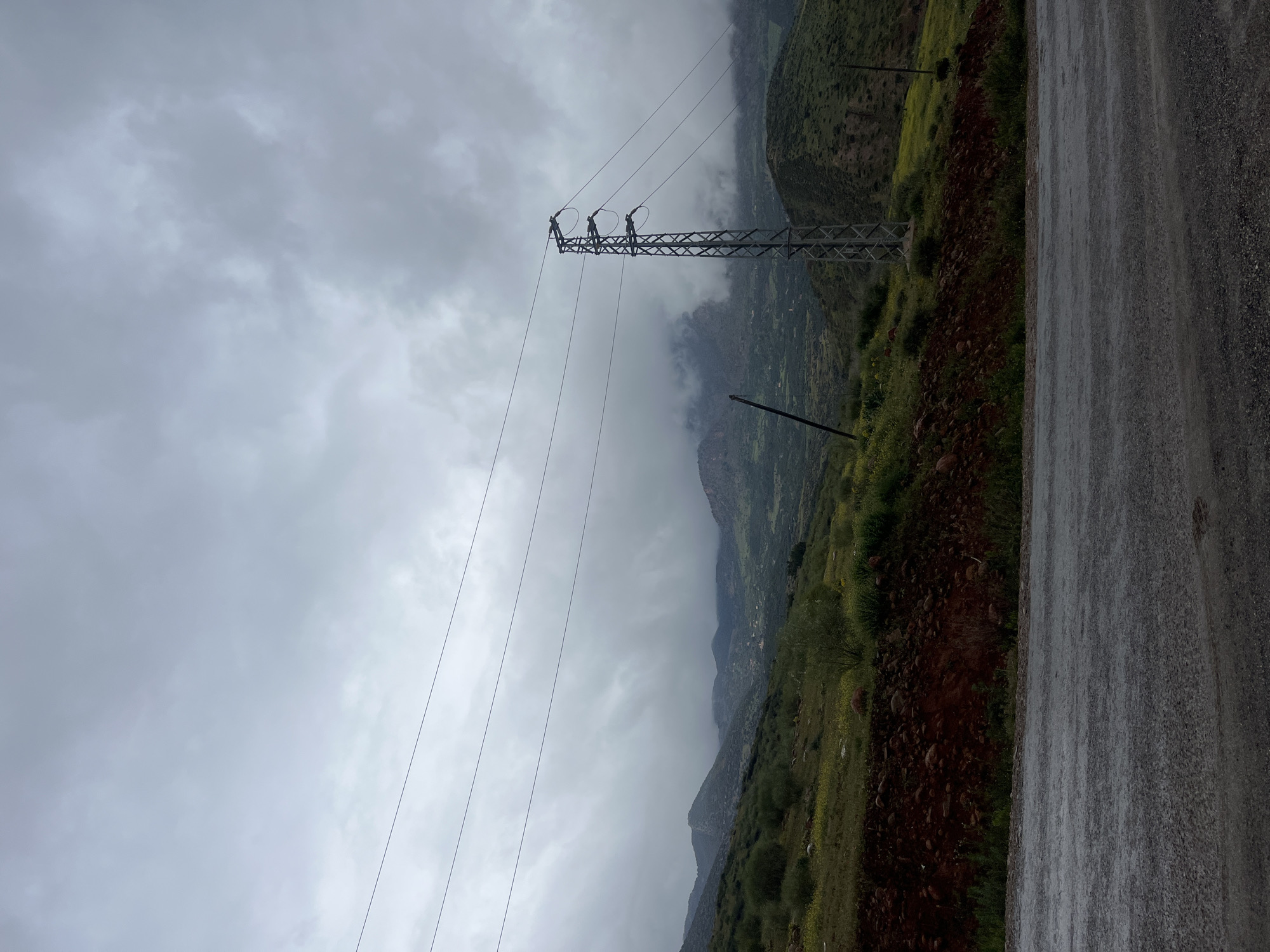 The image shows a landscape near Ouaouizert, Morocco, under a cloudy sky. A metal power transmission tower stands prominently, with three individuals appearing to be working on the upper section. Power lines extend from the tower towards the left. The terrain features green vegetation on hillsides, with some rocky, reddish-brown soil visible. A road with a wet, textured surface is visible on the right side of the frame. Mist or low clouds are present in the background, obscuring the upper parts of the mountains.