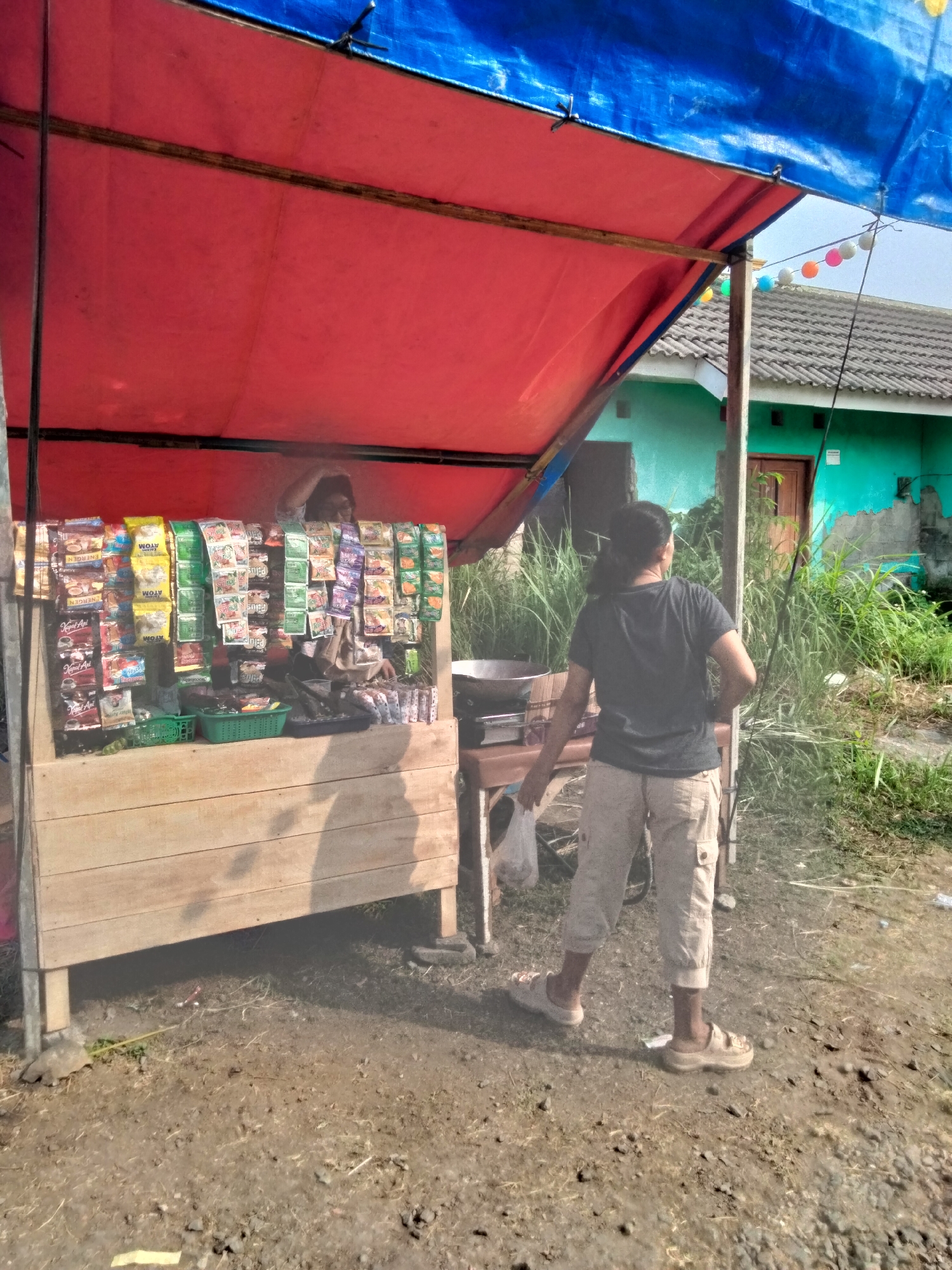 The image depicts an outdoor scene at a roadside stall in Cariu, Indonesia. A woman wearing a dark t-shirt and light-colored cargo pants stands behind a wooden stall displaying various packaged goods, likely snacks and beverages. The stall is covered by a red tarp and a blue tarp. In the background, a teal-colored building with a tiled roof is visible, partially obscured by tall grass. Decorative string lights with colorful bulbs are also visible above the stall. The ground is dirt with scattered debris. The woman appears to be interacting with the stall or tending to it. The overall impression is that of a small, local business operating in a semi-rural setting.