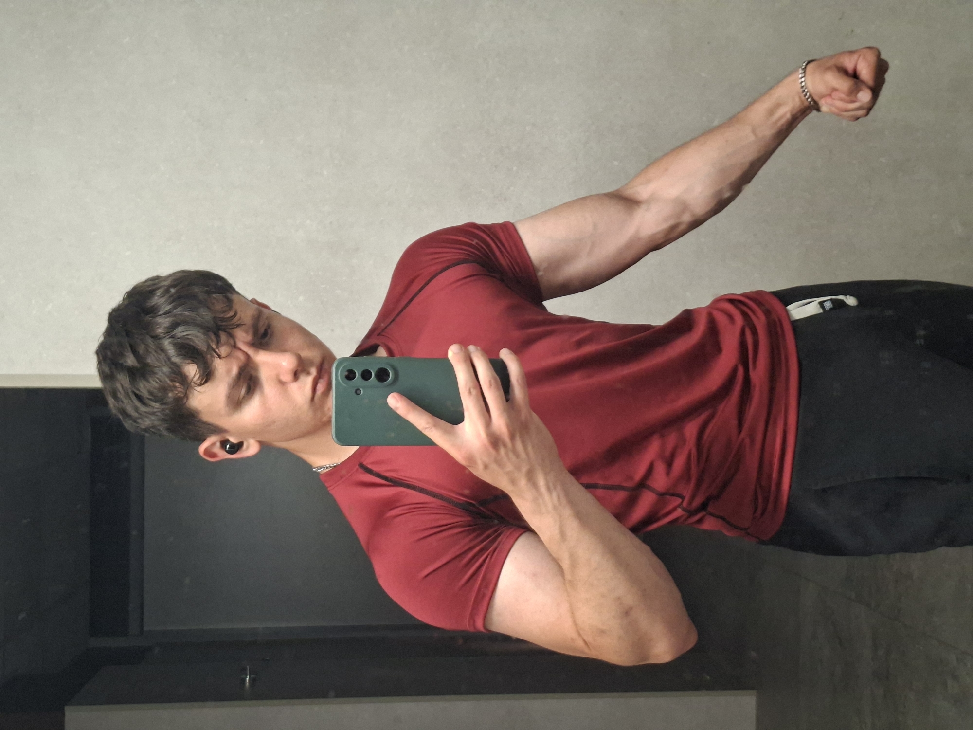 The image, taken in Plovdiv, Bulgaria, depicts a young man capturing a mirror selfie indoors. The subject appears to be in his late teens or early twenties, with short, dark, wavy hair and an earbud in his left ear. He is wearing a dark red athletic t-shirt with subtle black stitching and dark-colored athletic shorts or pants. A silver chain necklace is visible around his neck, and he wears a silver chain bracelet on his right wrist. His arms show significant muscle definition and visible veins, particularly the flexed right arm, which is raised with a clenched fist. He holds a dark green smartphone, featuring multiple cameras, in his left hand, pointing it towards the mirror to take the picture. His expression features slightly puckered lips, hinting at a playful or confident mood.
The background consists of light grey walls. A large, dark reflective surface, likely a mirror, occupies the space behind the man. On his left hip/waist, a white fitness tracker or band is attached to his shorts. In the lower part of the reflection, a dark, rectangular surface, possibly a counter or vanity with a faucet, is partially visible, suggesting the location could be a bathroom, gym changing room, or similar indoor setting with a large mirror. There is no discernible text visible in the image.
