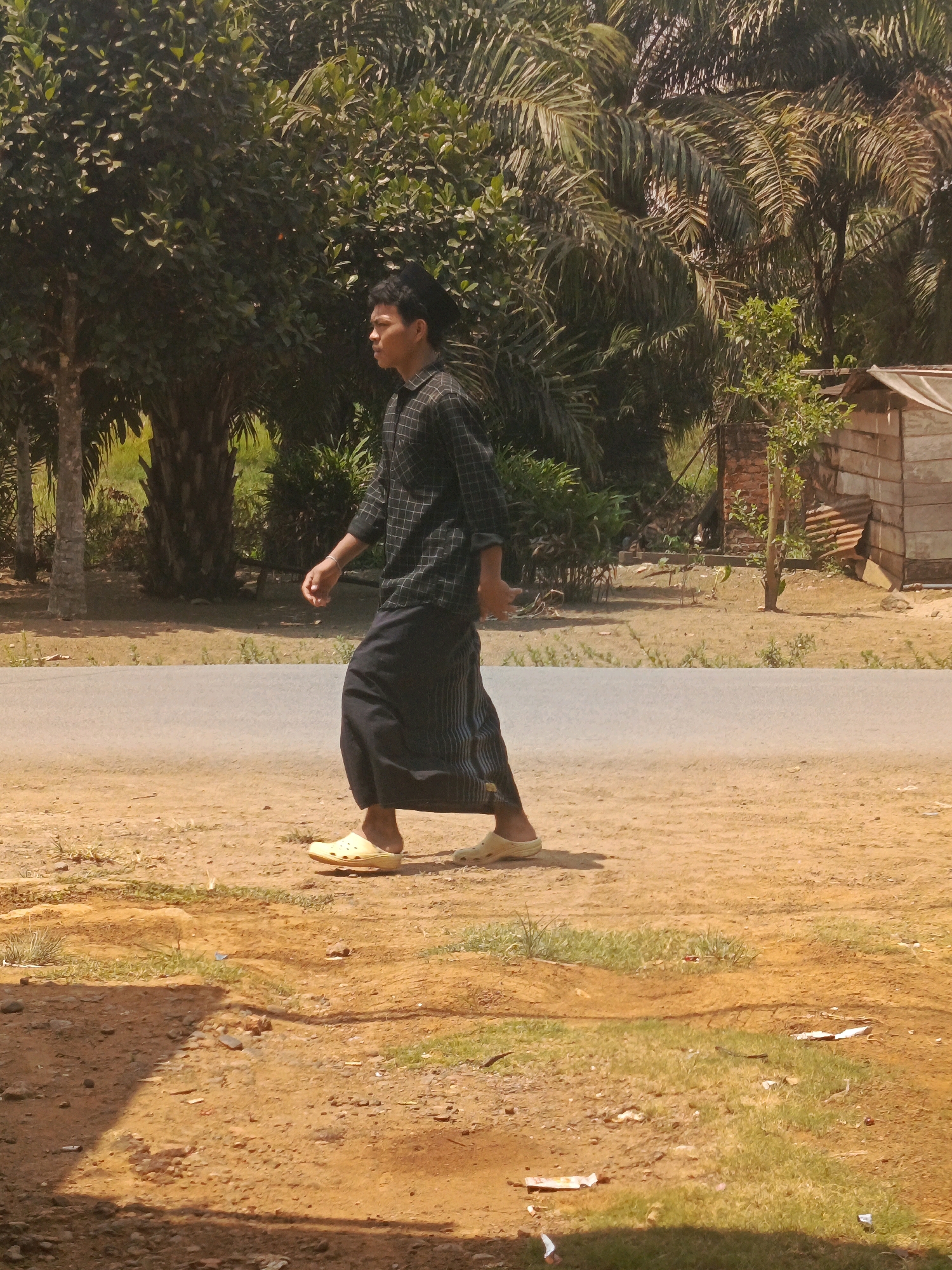 In the city of Megang Sakti I, Indonesia, a young man walks along a dirt road beside a paved asphalt road. He is wearing a black and white checkered long-sleeved shirt and a black sarong that reaches his ankles. His feet are clad in light yellow slip-on sandals. The background is dominated by lush green trees, including palm trees, and some vegetation. To the right, a simple wooden structure with a corrugated metal roof is visible. The ground is a mix of dry dirt, patches of grass, and scattered debris. The scene is illuminated by bright sunlight, casting shadows on the ground.