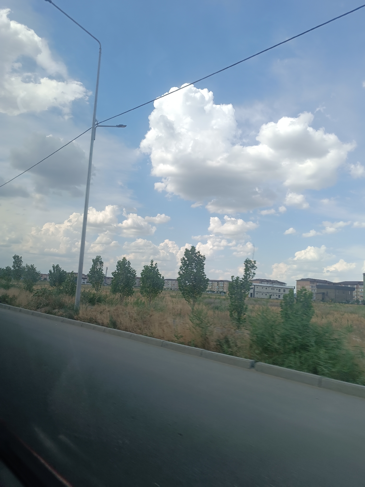 A scenic view captures a street in Taraz city, Kazakhstan, on a clear day with scattered white clouds against a vibrant blue sky. The image is dominated by a tall, light-colored street lamp positioned on the left side, with its silhouette cutting across the sky. A thin wire runs horizontally across the upper portion of the frame, seemingly connected to the lamp.
In the midground, a strip of dry, golden grass runs along the side of a paved road, suggesting a somewhat arid or dry climate. A row of green trees with full foliage stands interspersed with the dry grass, creating a visual division between the road and the distant buildings. Beyond the trees, several apartment buildings with light-colored facades and dark-tiled roofs are visible, hinting at a residential area. The architecture of these buildings appears modern and uniform.
The foreground features the asphalt road, which occupies the bottom half of the image. The road appears smooth and well-maintained, with a grey curb running along its edge. The angle of the shot suggests the photograph was taken from a moving vehicle, possibly looking slightly downwards towards the road. The overall impression is one of a quiet, perhaps developing, urban or suburban environment bathed in bright daylight. No people are visible in the frame, and no text is apparent. The weather is pleasant, indicative of a sunny day.