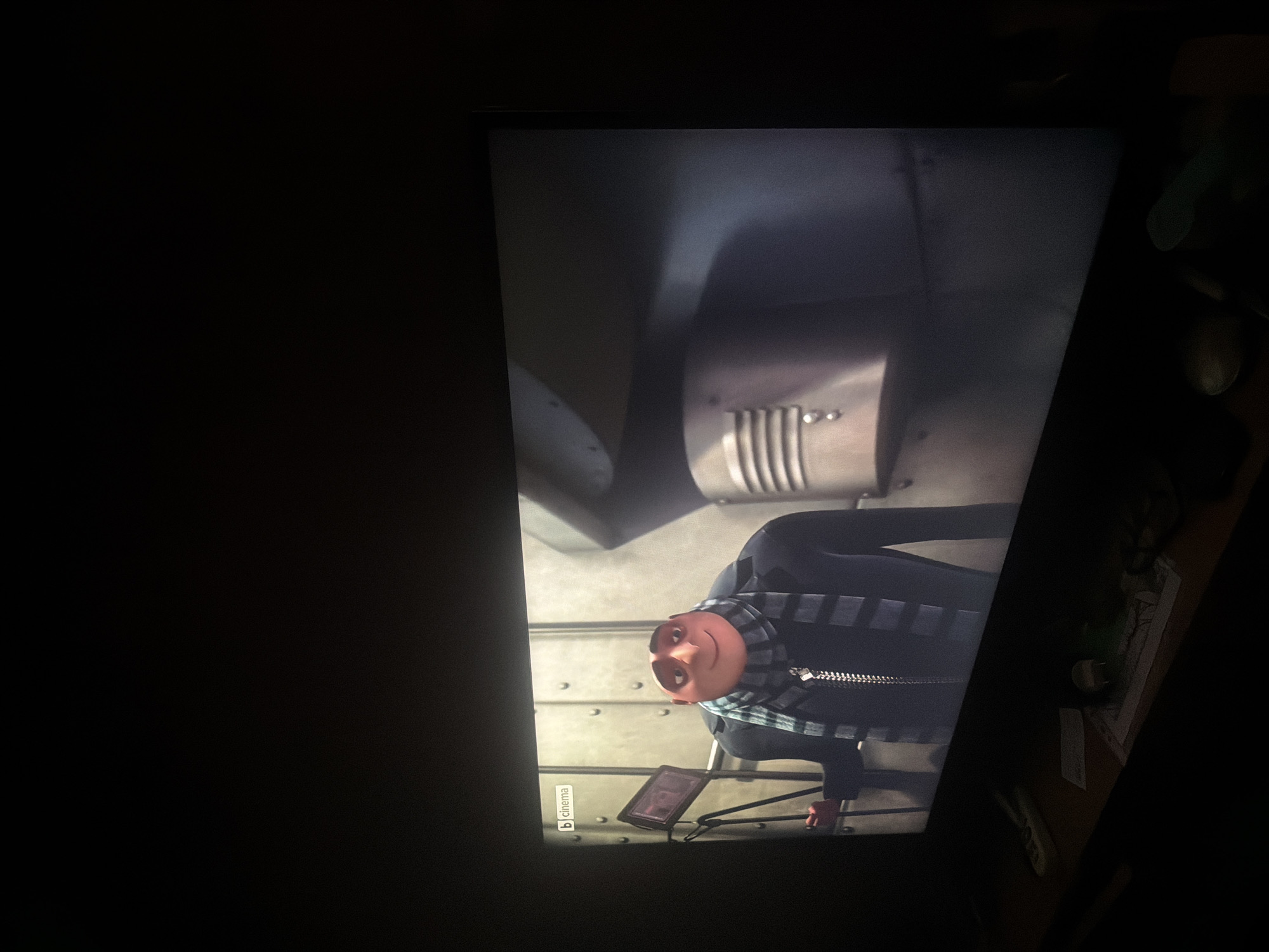 The image shows a television screen displaying the character Gru from the "Despicable Me" franchise. Gru is depicted inside a metallic, industrial-looking setting. A small "b cinema" logo is visible in the lower left corner of the screen. The room is dark, with the television being the primary light source. A desk and some miscellaneous objects are partially visible on the right side of the television screen.