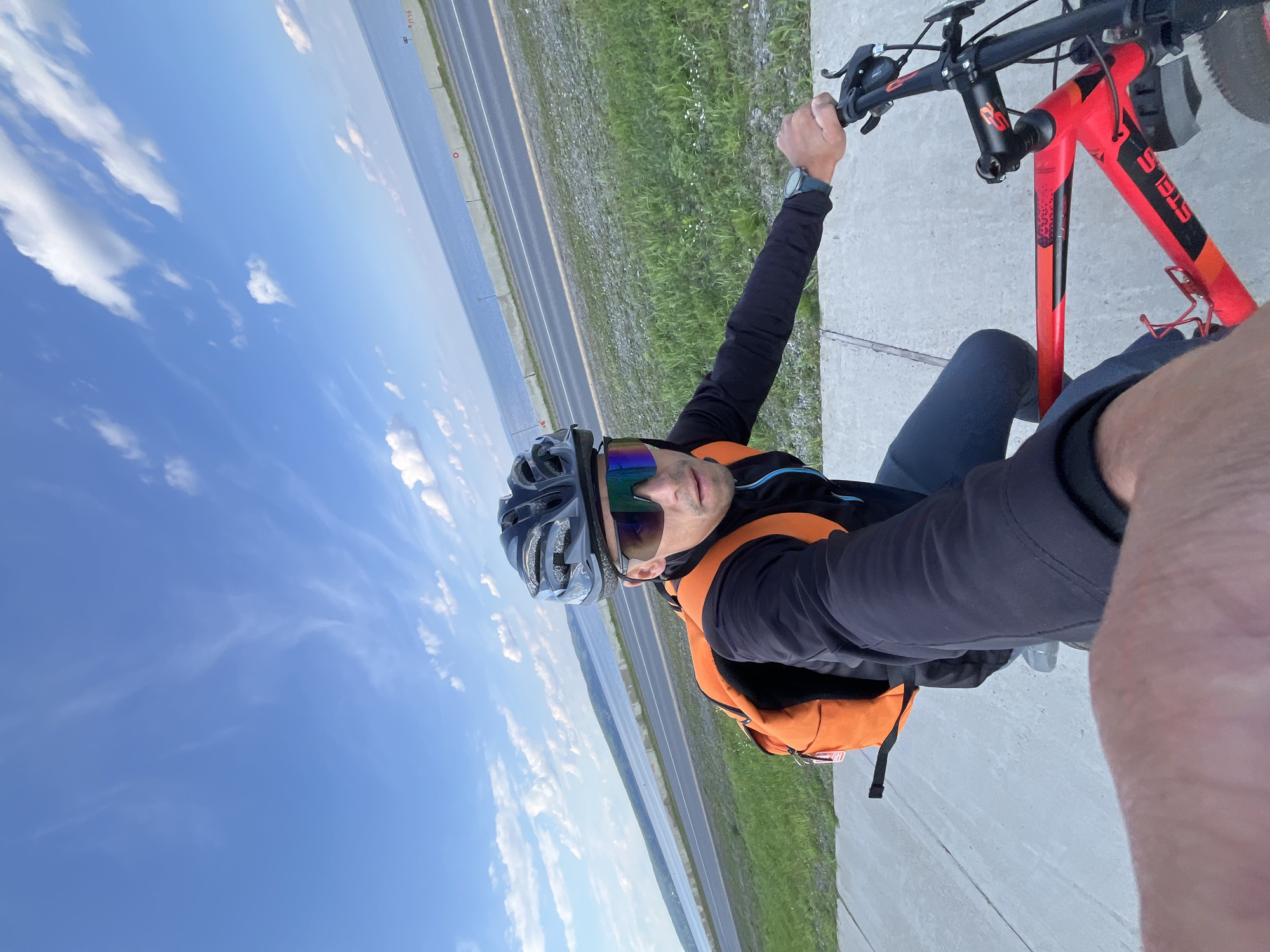 A selfie video captures a person on a red and black Stels bicycle, wearing a black helmet, dark sunglasses, a black jacket with blue accents, and grey pants. The person has an orange backpack. They are riding on a concrete path next to a grassy area, with a road, a water body, and a distant shoreline visible. The scene is set against a bright blue sky with scattered white clouds, suggesting it is daytime. The location appears to be near the Kama River in Chaikovsky, Russia.