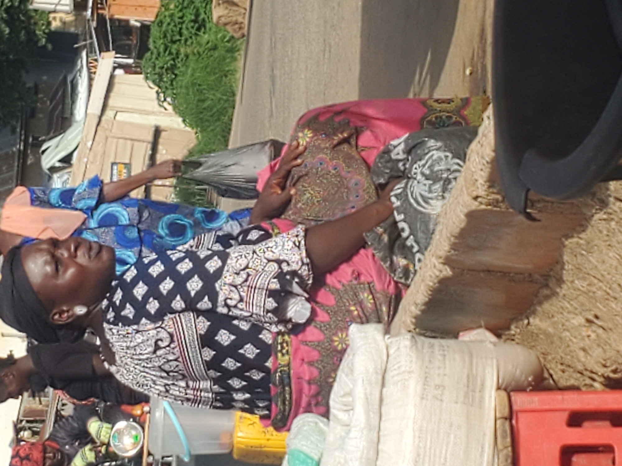 A medium shot shows a woman sitting on a curb in Gwong, Nigeria. She wears a black headscarf, a black and white patterned blouse with wide sleeves, and a pink skirt with gold trim. Her left hand rests on her lap, and her right hand is raised slightly. In front of her are several large white sacks, a red plastic crate, and a black trash can. Behind her, a person in a blue and orange patterned outfit carries a black plastic bag. In the background, a small wooden structure and lush green foliage are visible, suggesting a marketplace or roadside setting. The scene is likely captured during daylight hours.