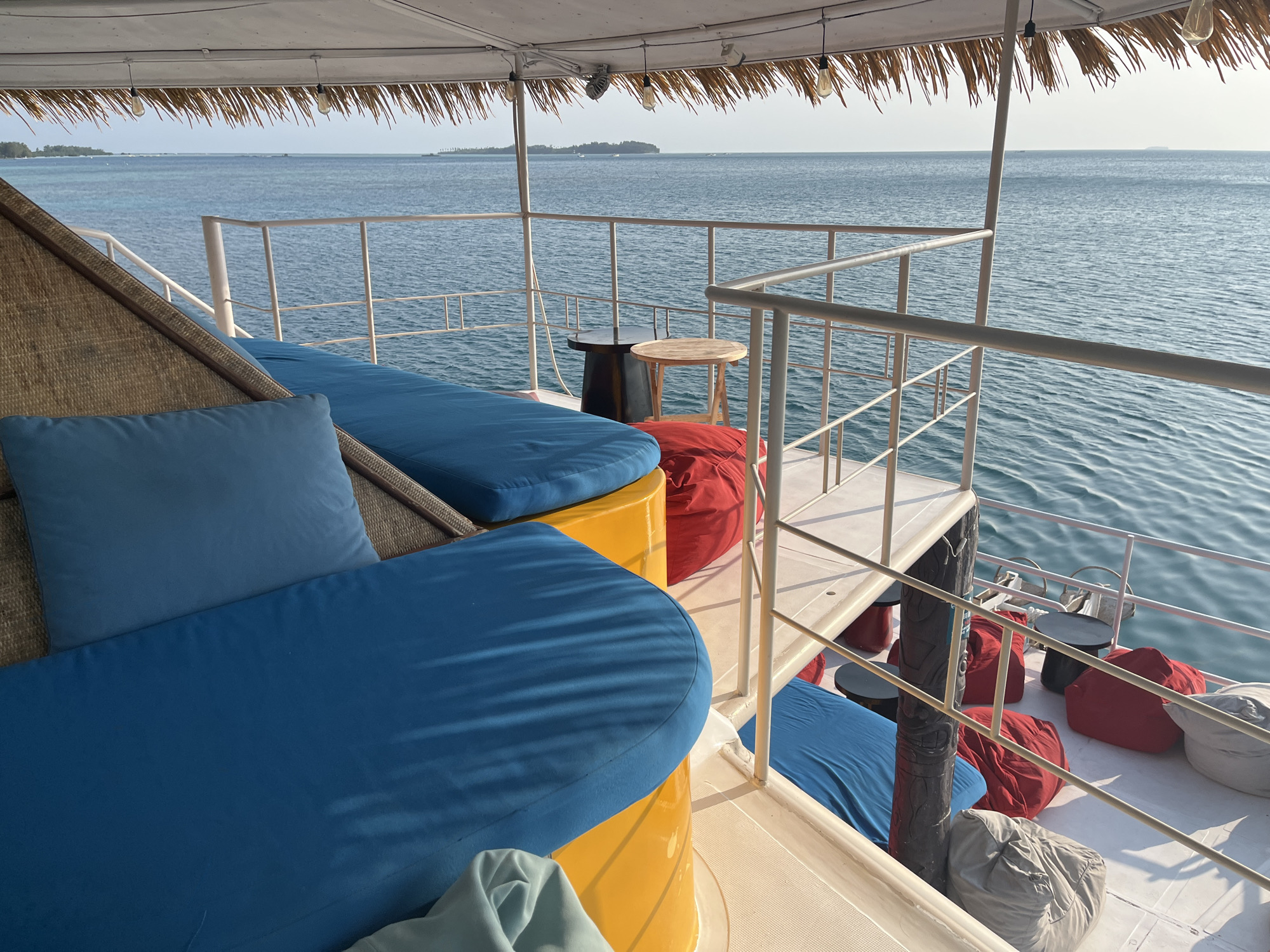 This image captures a scenic view from the deck of a boat in Karimunjawa, Indonesia. The deck is furnished with several blue cushioned lounge chairs and red and gray beanbag chairs, suggesting a comfortable seating area for passengers. In the background, the calm, blue ocean stretches towards the horizon, with a few distant islands visible. The sky is clear and bright, indicating a pleasant day. A thatched roof provides shade over part of the deck, and decorative string lights are suspended from it. The boat appears to be docked or moving slowly, as the water is relatively still. The overall scene conveys a sense of relaxation and leisure, typical of a tropical vacation destination.