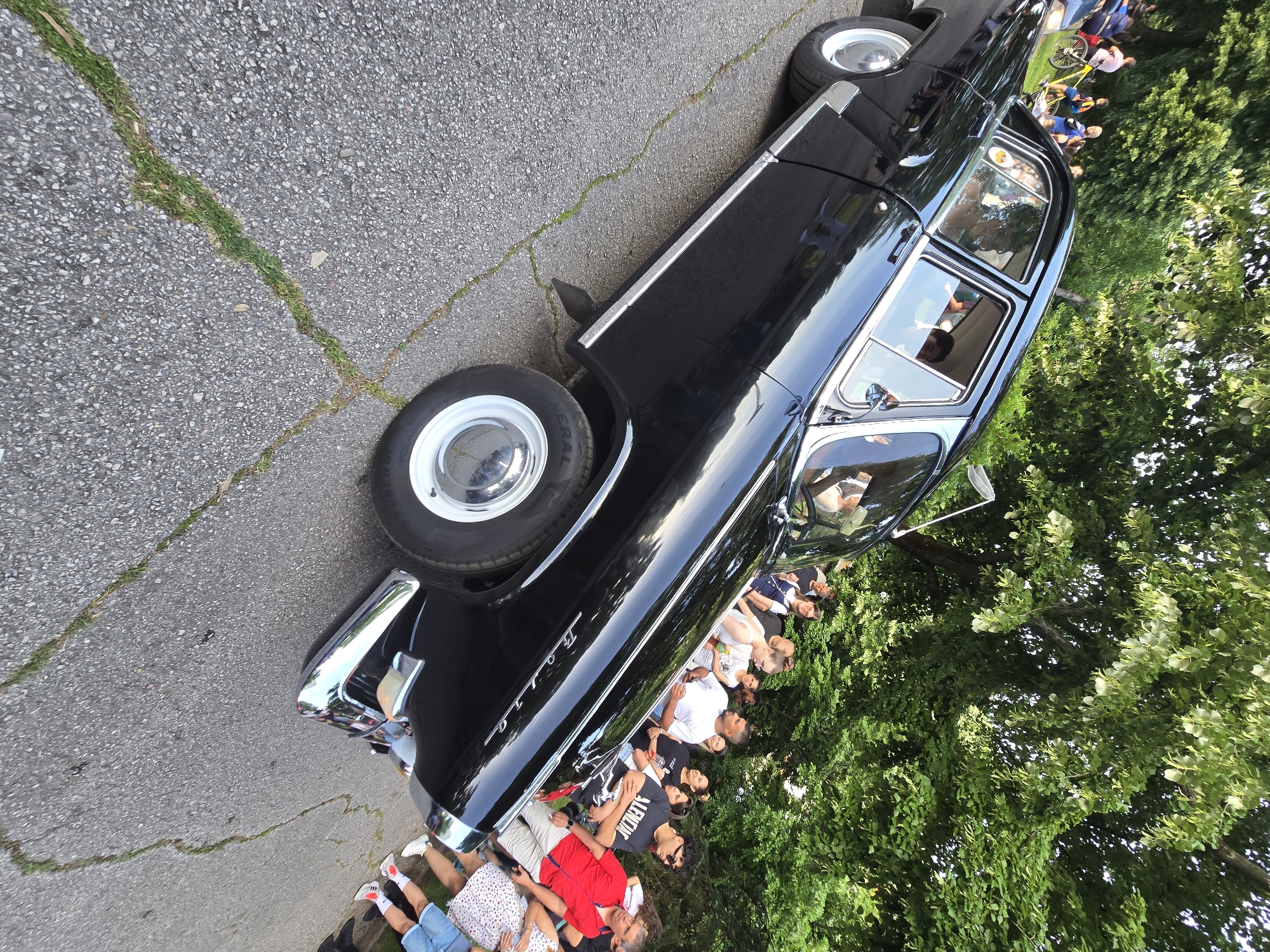 The image shows a black vintage car parked on an asphalt road with cracks filled with grass. A crowd of people are gathered on the grass to the right of the car. The background consists of green trees. The car is a "Volga" model.