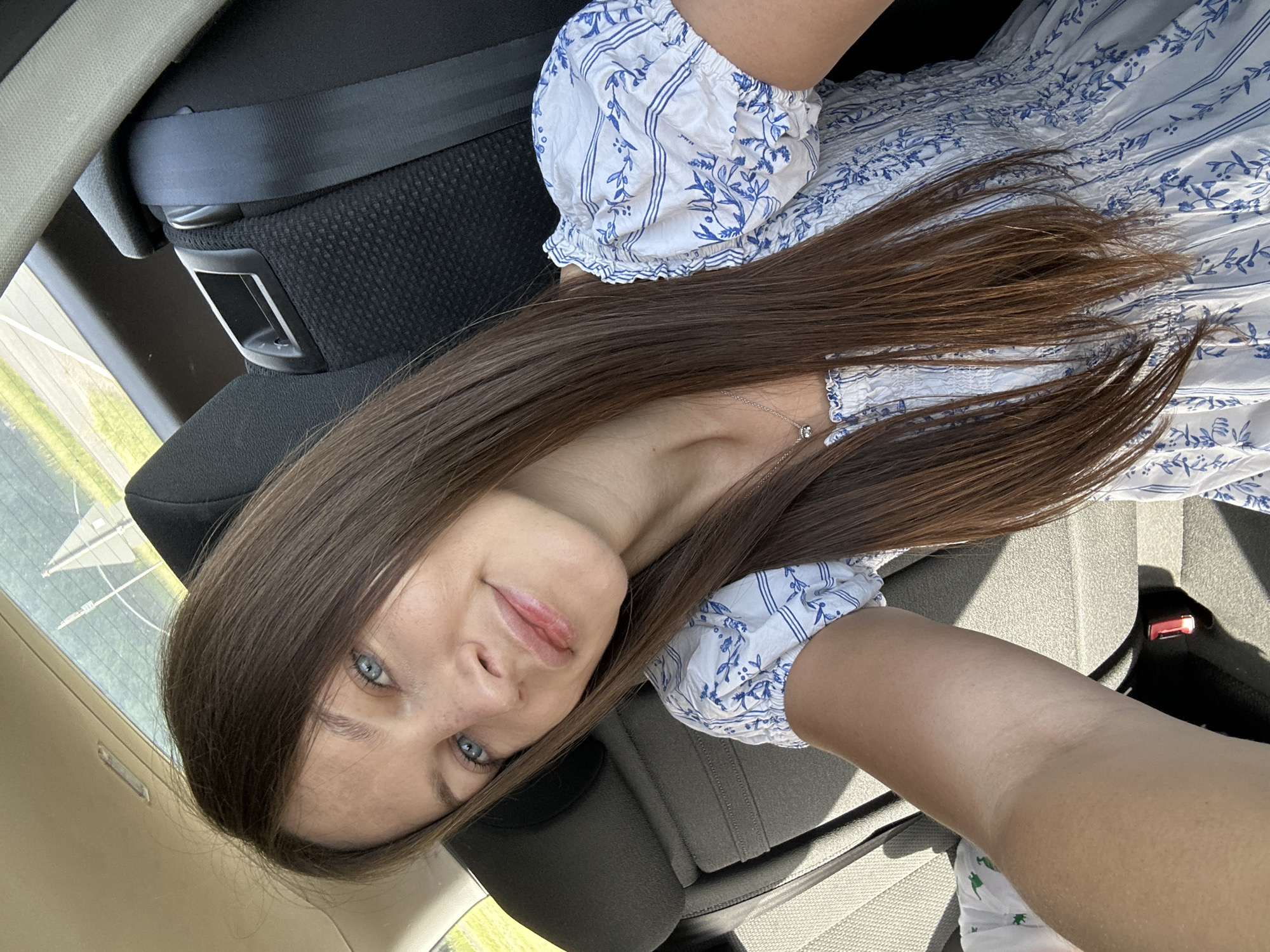 A woman is in the back seat of a car. The car is presumably in Niederquembach, Germany, since that was specified for the location of the image. She is wearing a white and blue patterned dress. She is smiling slightly and looking at the camera. Her long brown hair is spread out on the car seat. She is wearing a necklace. The car's seatbelt is visible. The car's interior is various shades of black and grey. Outside the window is a view of a green field.
