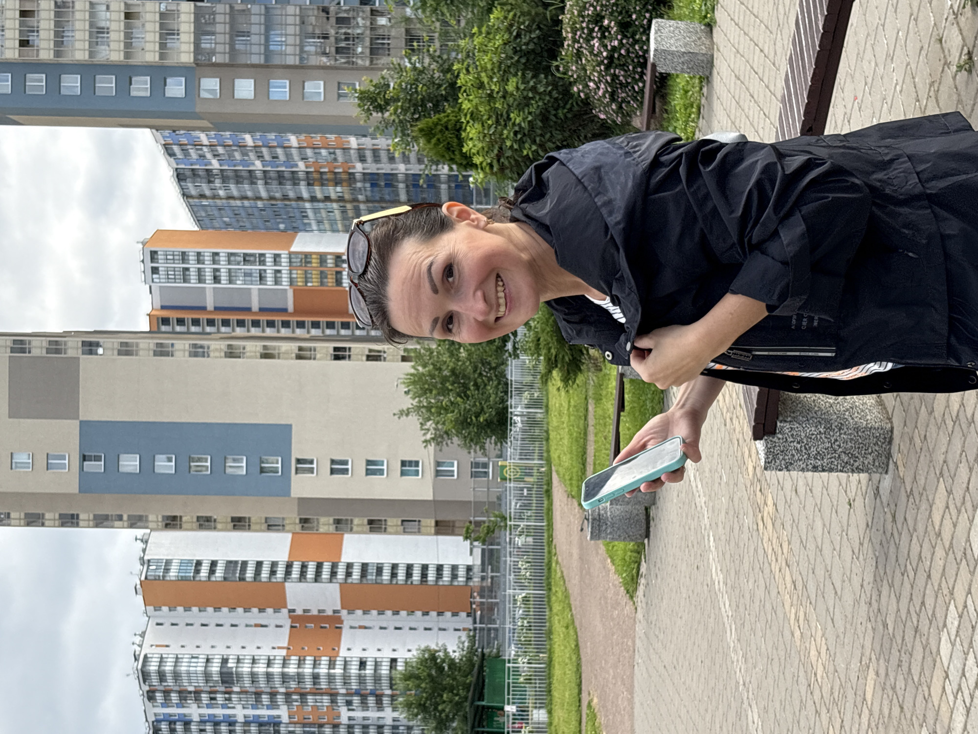 A woman is standing outside holding a smartphone. She is wearing a black jacket over a white shirt. Her hair is pulled back and she is smiling at the camera. In the background, there are several tall buildings, some of which are brightly colored. The buildings appear to be residential apartments, and the overall scene suggests an urban environment in Saint Petersburg, Russia. The ground is paved with interlocking bricks. A park bench is visible to the right of the woman.