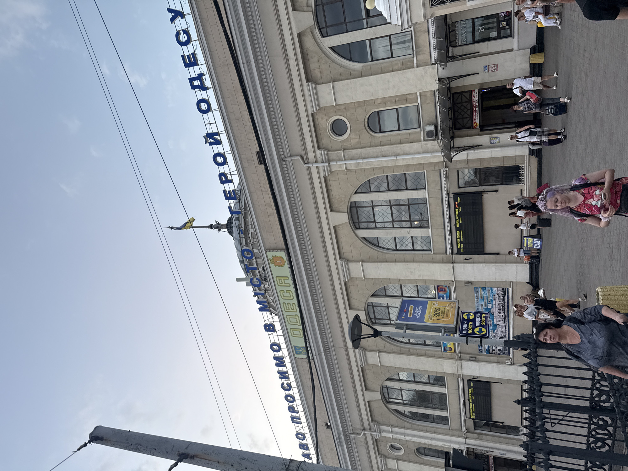 This is a vertical, eye-level shot of a street scene in Odesa, Ukraine. The foreground is dominated by a grey sky and a utility pole. The midground shows a building with arched windows and ornate architectural details. A large blue sign with Cyrillic text, "АВО ПРОСИМО В МІСТО ГЕРОЙ ОДЕСУ" ("Welcome to the Hero City of Odesa"), spans across the top of the building. A yellow and blue Ukrainian flag is visible on a pole attached to the building.
The background features a paved area with several people walking and standing. Some individuals are carrying luggage, suggesting a transportation hub or a busy tourist area. Signs on the building advertise "КАСИ" ("TICKETS"), "ТУАЛЕТ" ("TOILET"), and directional arrows for "ПІВНІЧ NORTH" ("NORTH") and "ПІВДЕНЬ SOUTH" ("SOUTH"). There are also advertisements for discounts and local services. The lighting suggests it's daytime, likely a bright, possibly warm day given the light clothing worn by some of the people. The overall mood appears bustling and active, indicative of a public space with people on the move.
