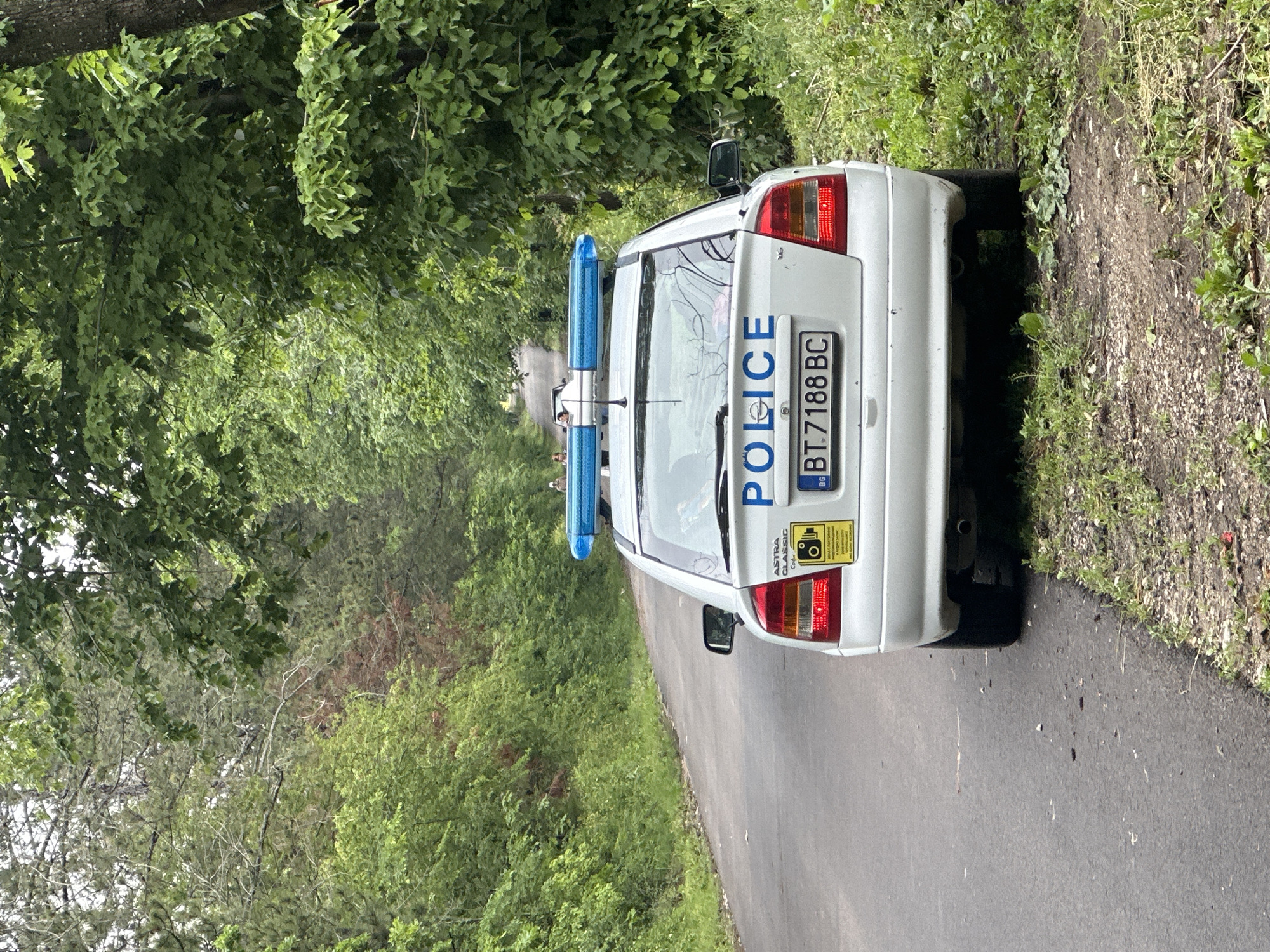A white Opel Astra Classic police car is parked on a narrow asphalt road, likely on the outskirts of a town or village. The rear of the car is facing the camera. The license plate reads "BT.7188 BC." The word "POLICE" is vertically printed on the car's trunk. Two blue emergency lights are mounted on the roof. The car is flanked by lush green vegetation on both sides, including trees and bushes. Based on the license plate code "BT," it's possible the scene is located in or around Veliko Tarnovo Province, Bulgaria. The presence of the Opel Astra Classic car could suggest the approximate year of this photo taken is mid-2000s. The exact location in Sheremetia, Bulgaria, cannot be determined without additional visual references.