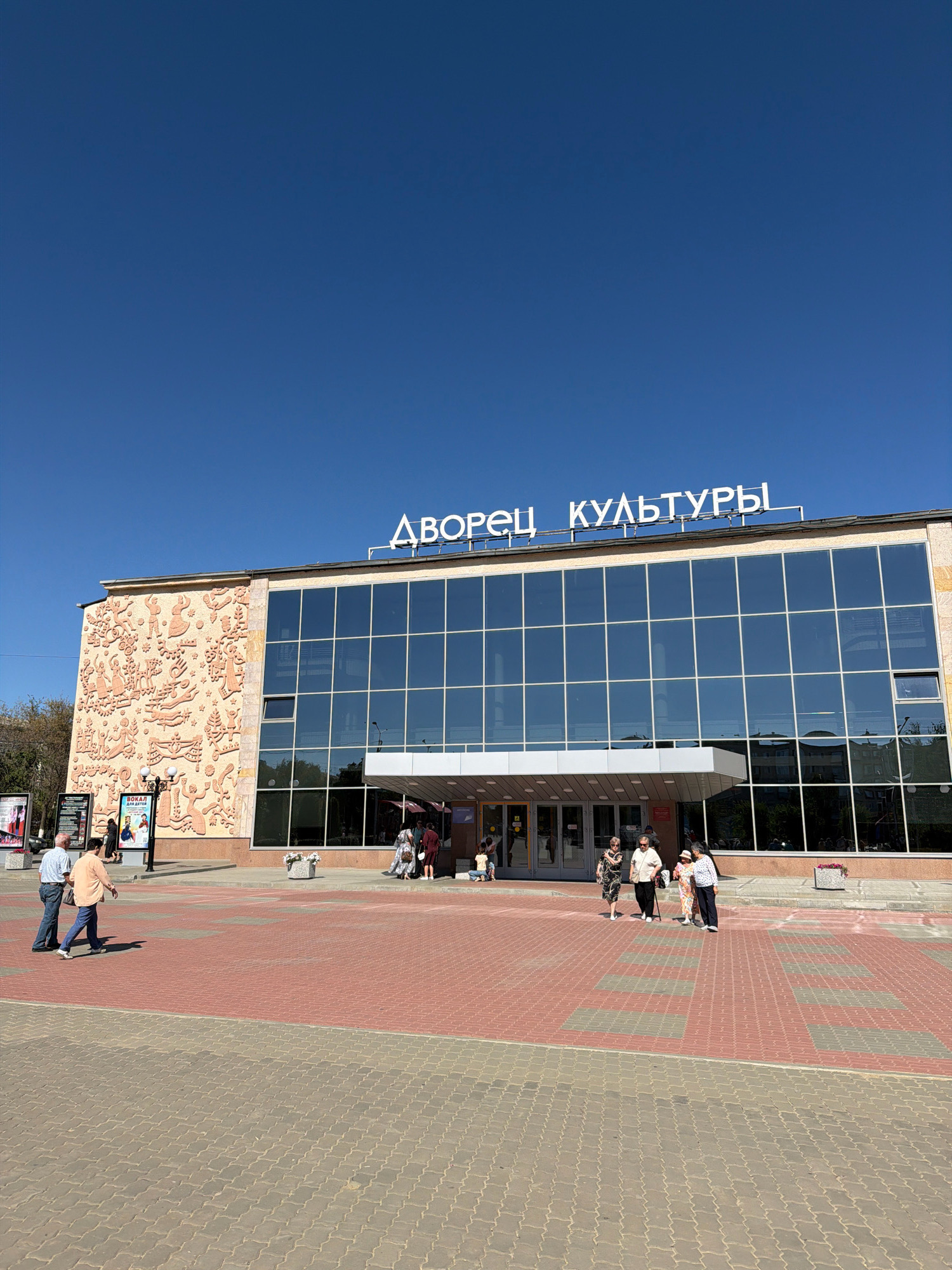 The photograph captures the Palace of Culture in Kamyshin, Russia, on a clear, sunny day. The building's facade is a mix of modern glass windows and an ornate, relief-decorated wall on the left. White, blocky lettering above the glass expanse spells out "ДВОРЕЦ КУЛЬТУРЫ" (Palace of Culture). The area in front of the building is paved with interlocking bricks, a pattern of red and gray blocks.
Several people are visible on the paved area. On the left side, two men in casual clothing are walking. Further towards the entrance of the building, a group of four individuals, appearing to be women, are strolling. Another person, a child, is visible sitting on the ground near the entrance. In the background, a poster displaying an advertisement is mounted on a stand. The sky above is a vibrant, unblemished blue.