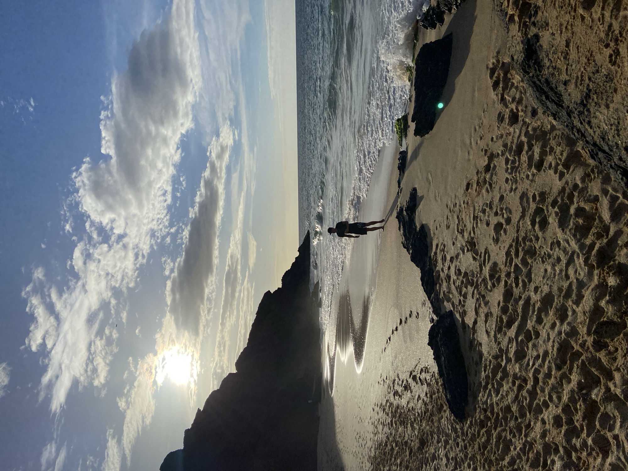 This image depicts a beach scene with a person standing on the sand near the water's edge. The sun is setting or rising, casting a warm glow and creating reflections on the wet sand. A dark, rocky cliff face is visible in the background on the left, while the ocean stretches out to the horizon on the right. The sand is textured with footprints and scattered dark rocks. The sky is partly cloudy, with the sun partially obscured by clouds. The location is not specified as Altagracia, Venezuela.