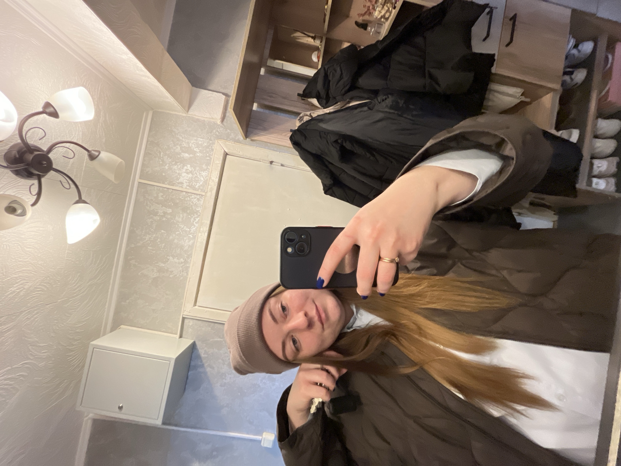 A woman wearing a brown beanie and a brown jacket is taking a selfie in a room with textured wallpaper and a white cabinet. The room also contains a black puffer jacket hanging near a wooden shelf or closet with clothing and shoes. The lighting is provided by a multi-bulb ceiling fixture. The location is Razdolye, Russia.