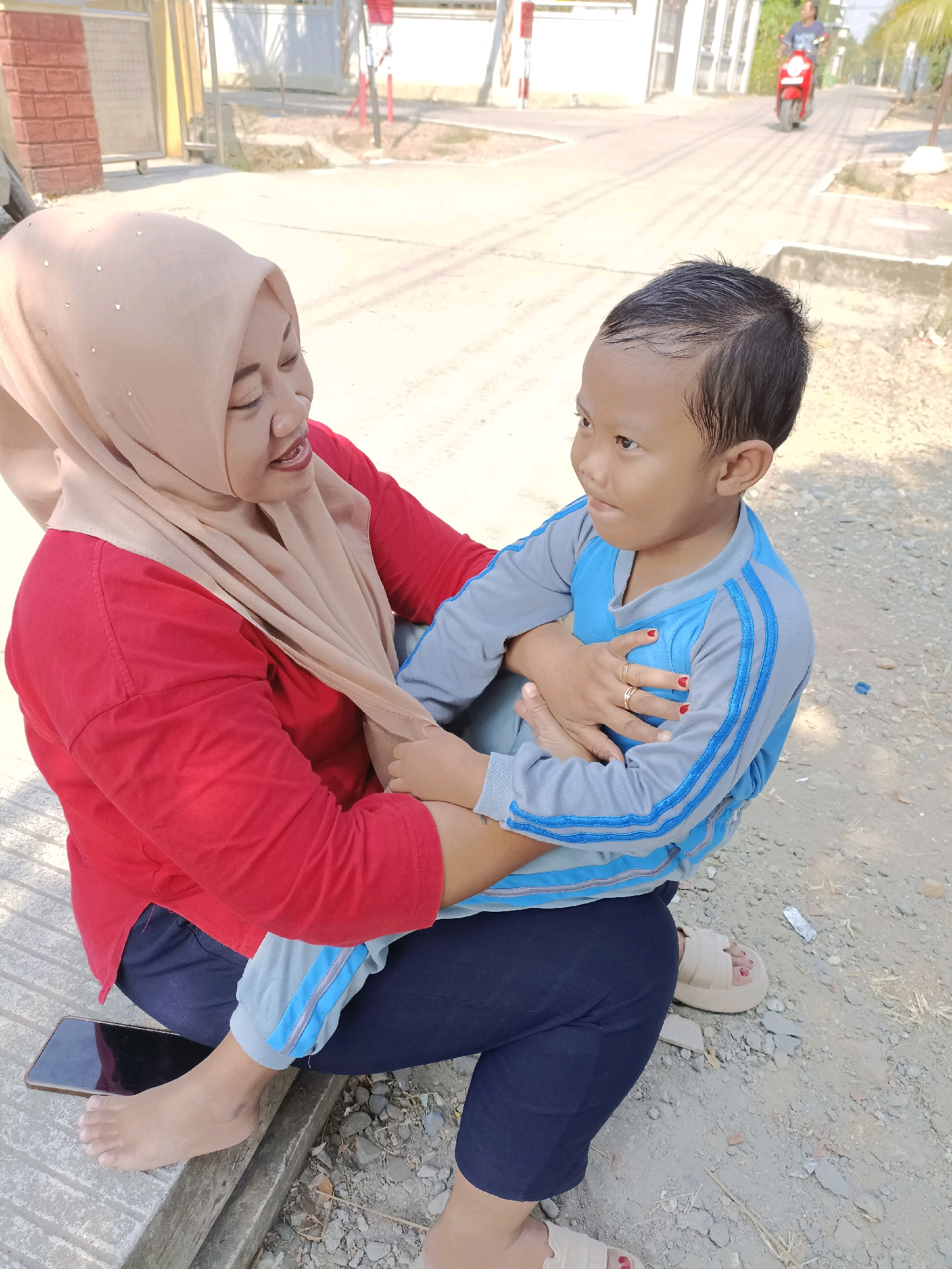 A woman wearing a hijab and a red shirt is sitting on a wooden bench, holding a young child on her lap. The child is wearing a blue and gray tracksuit. A smartphone is placed on the bench next to the woman's feet. The background shows a street with a person riding a red motorcycle in the distance. The scene appears to be outdoors in a residential area of Cangkring, Rembang, Indonesia, on a sunny day.