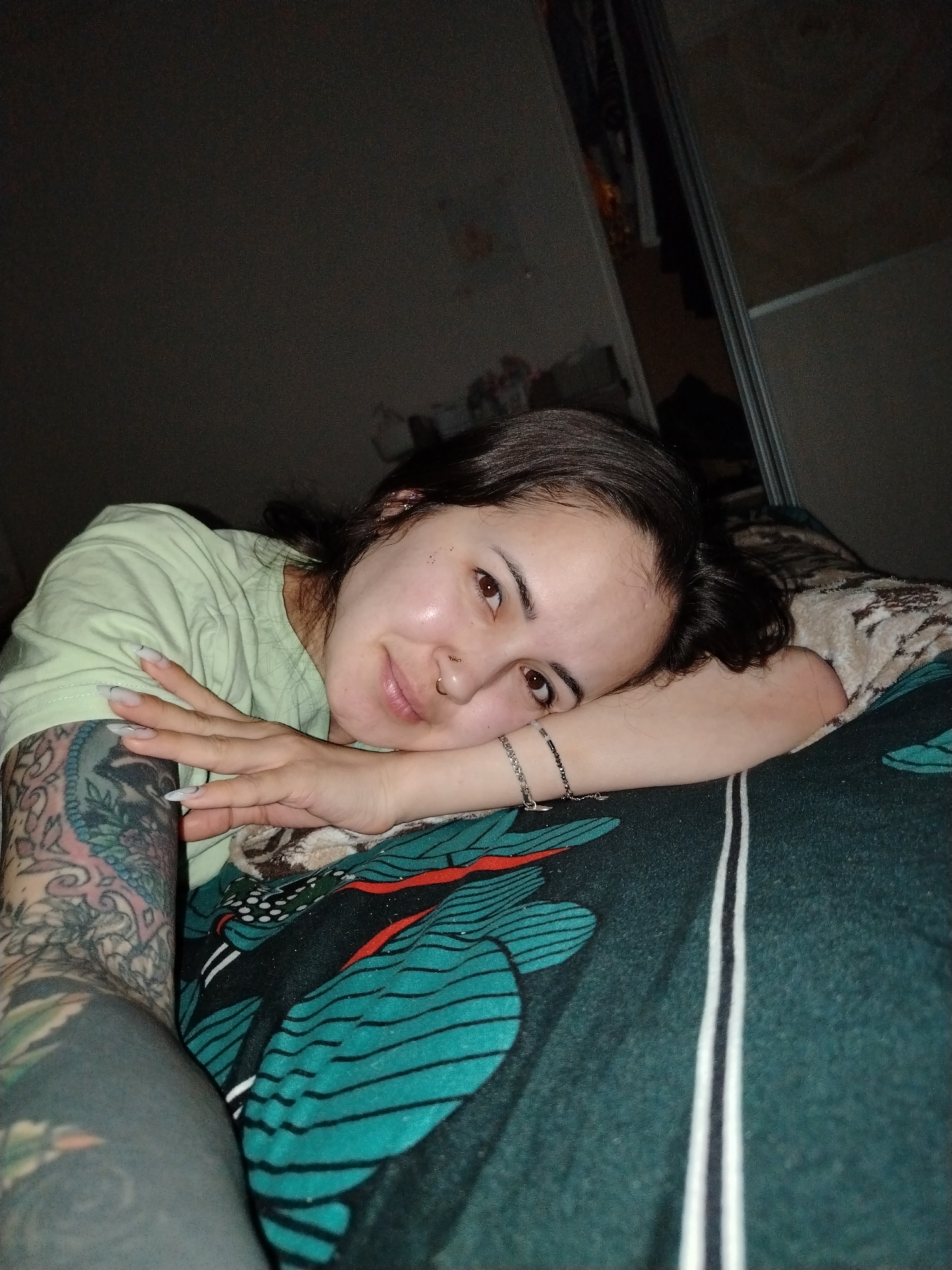 A woman with dark hair and fair skin is lying on a bed. She is wearing a light green t-shirt and has multiple tattoos on her left arm. She is smiling slightly and has a nose ring. She is wearing bracelets on her right wrist. The bed is covered with a patterned blanket, including what appear to be representations of green feathers and a white and dark patterned pillow. In the background is a dimly lit room with what appears to be a closet. There is no information available to ascertain the location in Noviy, Russia.