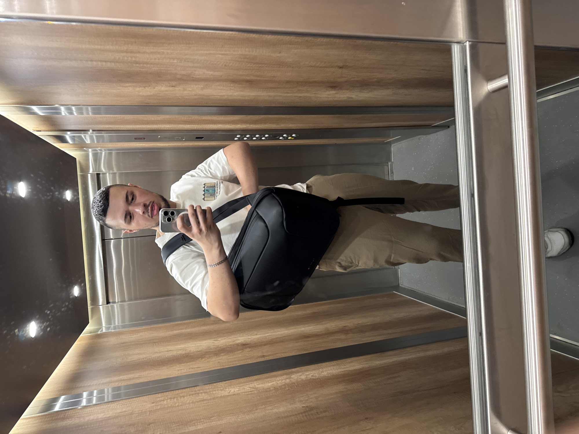 A man is taking a selfie in an elevator. He is wearing a white t-shirt with a design, tan pants, and white sneakers. He has a black backpack slung over his shoulder. The elevator has wood-paneled walls, a gray floor, and stainless steel doors. There are buttons visible on one wall. The location is likely Sofia, Bulgaria, as there is nothing to explicitly exclude it as a possible location for this image.