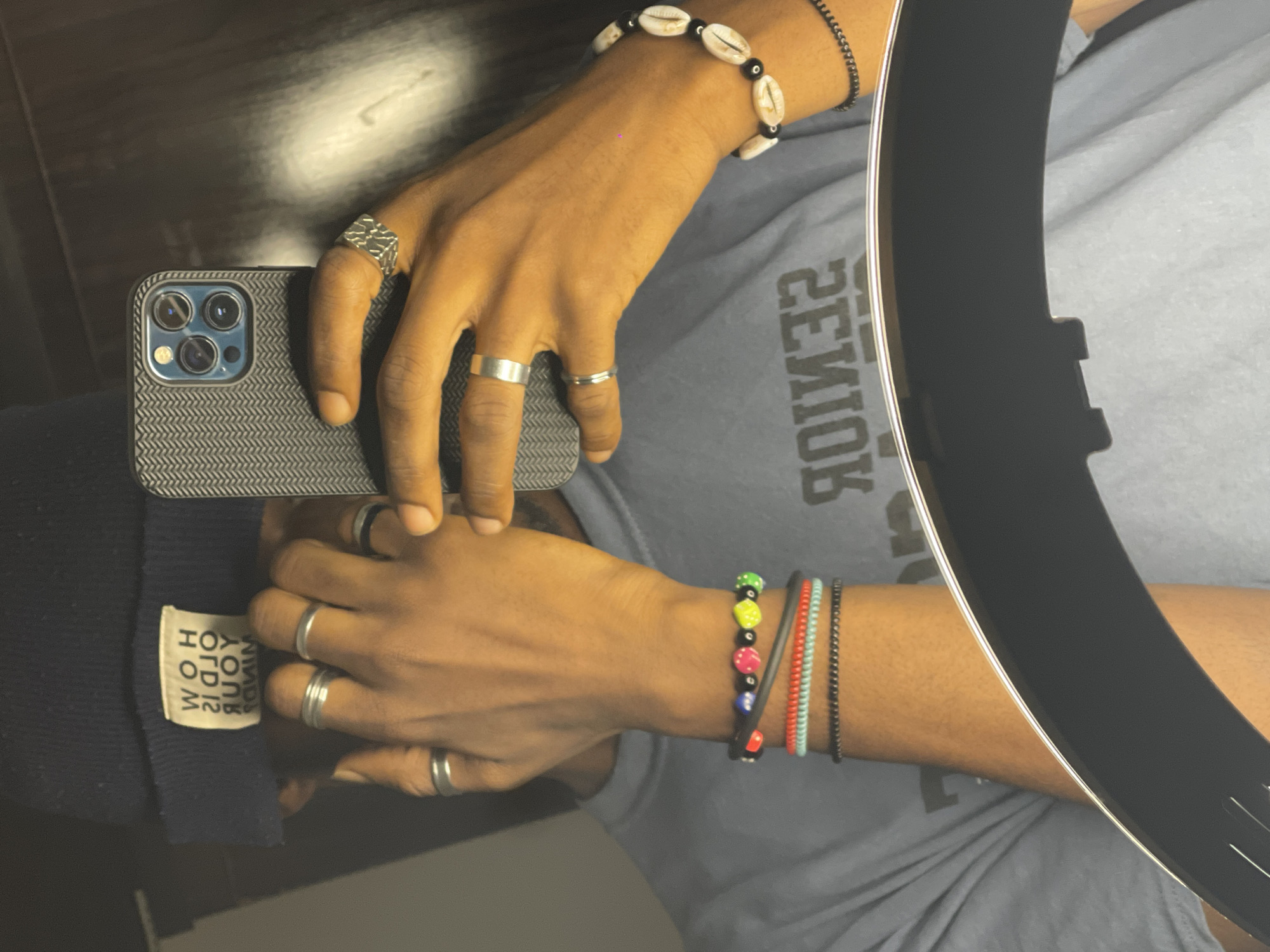 A close-up, waist-up shot shows a person holding a smartphone to take a mirror selfie. The person is wearing a dark blue beanie, a grey t-shirt, and several bracelets on their left wrist. The bracelets include a black and red beaded bracelet with colorful circular beads, a red cord bracelet, a light blue cord bracelet, and a black beaded bracelet. The person also wears multiple silver rings on their fingers, including a band ring on the index finger, a thicker band ring on the middle finger, and a patterned ring on the ring finger of their right hand. The smartphone, a silver iPhone, is held in the person's right hand, its patterned grey case clearly visible. In the background, a large ring light is partially visible in the foreground. The text on the t-shirt, in a mirrored font, reads "SENIOR". The image appears to be taken indoors.