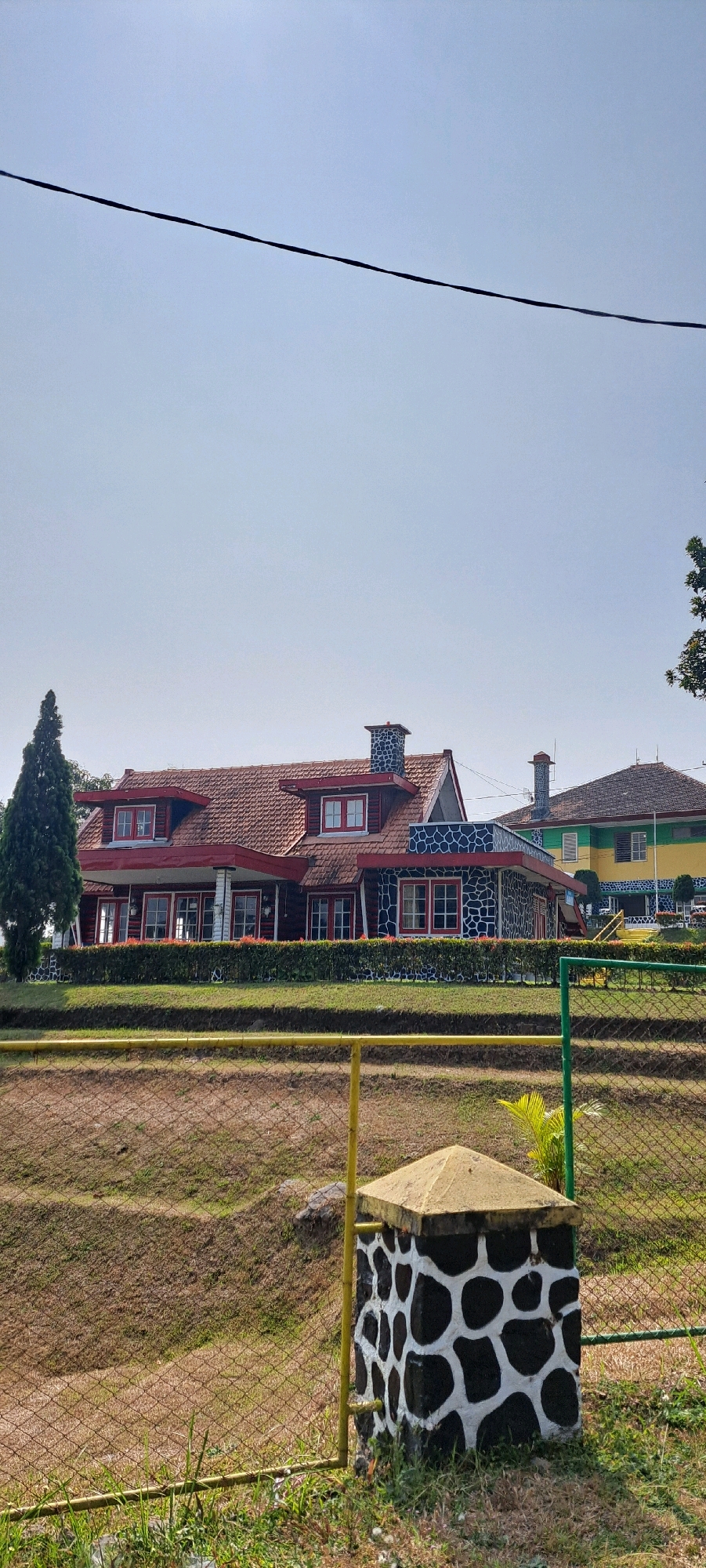 The image captures a view of a large, multi-story building with a prominent red tile roof and red-trimmed windows. The building's facade features sections with a dark gray stone pattern with white irregular shapes. A tall, dark green evergreen tree stands to the left of the building. In the background, to the right, another building is visible with a yellow and green facade and a brown roof. A wire stretches across the upper part of the frame against a clear, pale blue sky.
In the foreground, a chain-link fence with yellow and green poles encloses a grassy embankment. A decorative stone pillar with a black and white spotted pattern and a yellow cap stands near the fence. The ground in the immediate foreground is dry and covered with sparse vegetation. The scene appears to be set in an outdoor environment with a clear daytime sky. The location is the Pintu Langit Sky Gate tourist spot in Prigen, Indonesia.