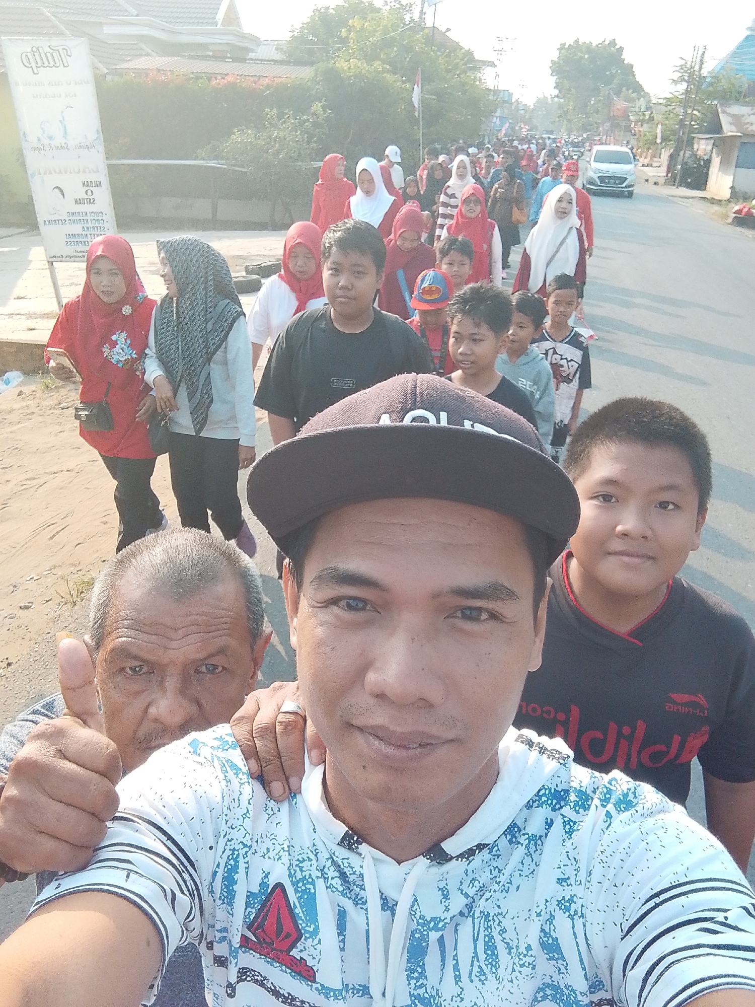 In Kias, Indonesia, a group of people are walking down a paved road. In the foreground, two men are visible. The man on the left, an older gentleman with graying hair and a mustache, is giving a thumbs-up gesture. He is wearing a gray shirt. Next to him, a younger man with a dark baseball cap is taking a selfie. He is wearing a white and blue patterned shirt. Behind them, a crowd of people, including men, women, and children, are walking in the same direction. Some of the women are wearing hijabs. A white car is visible in the distance on the road. The environment appears to be an urban or suburban area, with buildings and trees lining the street. A sign with text is also visible on the left side of the image. The lighting suggests it is daytime.