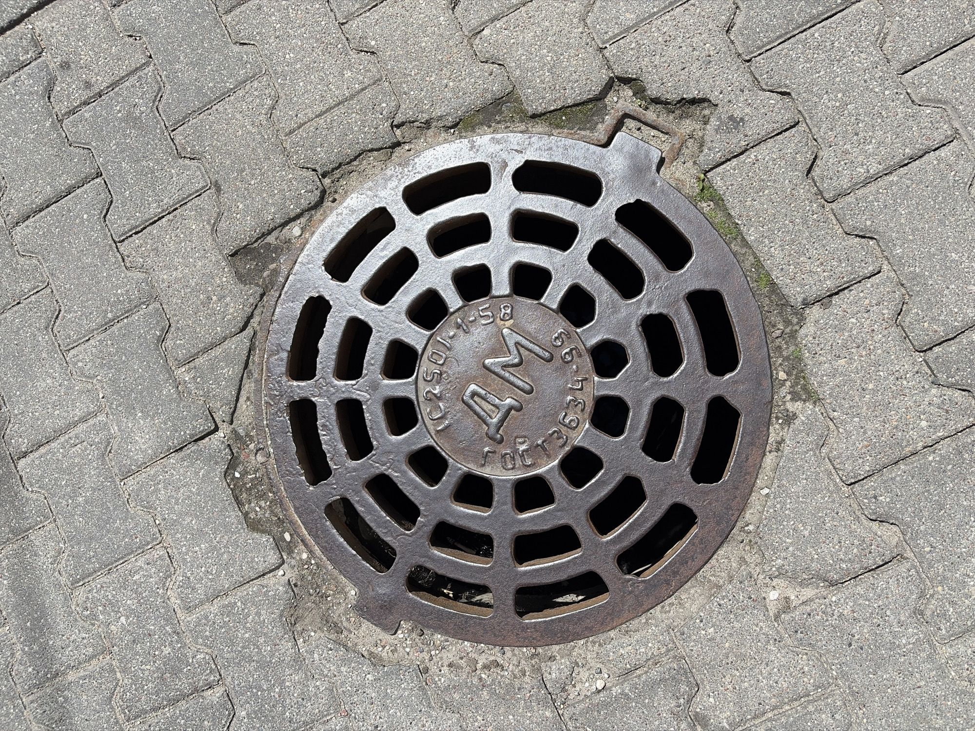 A top-down shot of a round metal manhole cover set into a paved sidewalk. The manhole cover has a grid pattern of openings and a circular emblem in the center. The emblem features the letters "AM" prominently, surrounded by smaller text and numbers that are partially obscured. The surrounding paving stones are interlocking and gray. The lighting suggests it is daytime, with strong shadows cast by the manhole cover.