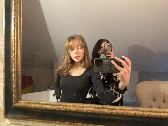 The image depicts a mirror selfie taken by two individuals in what appears to be a domestic interior. The location in the city of Unknown, Unknown cannot be determined.
The scene shows the reflection of two young adult females in a large, rectangular mirror with an ornate, dark gold/bronze frame.
One female (left foreground) has long, light brown hair with bangs, and wears a black, long-sleeved, scoop-neck top. Her right hand (viewer's left) rests near her hip, and her gaze is directed slightly to her left, with lips slightly parted.
The second female (right background, taking the photo) has dark, wavy hair. Her face is largely obscured by a light gold/silver smartphone she holds up. Her right arm and hand (viewer's right) are prominent, holding a dark-colored smartphone with a textured "pop-it" style case, aimed at the mirror to capture the image. Her fingernails are manicured with white tips, and a black scrunchie is on her right wrist. She wears a black top, with a segment of a black and white patterned garment visible on her upper torso.
The environment reflected in the mirror includes light-colored walls, one with a subtle textured pattern on the far left. A sloped ceiling is visible in the upper portion, suggesting an attic or mansard room. On the left side of the reflection, a white bathtub is partially visible, alongside a red fabric item (possibly a towel) draped over an unseen object, a white dispenser bottle, and a partial yellow object. On the right side, a section of a cream or beige upholstered armchair with tufted detailing is visible.