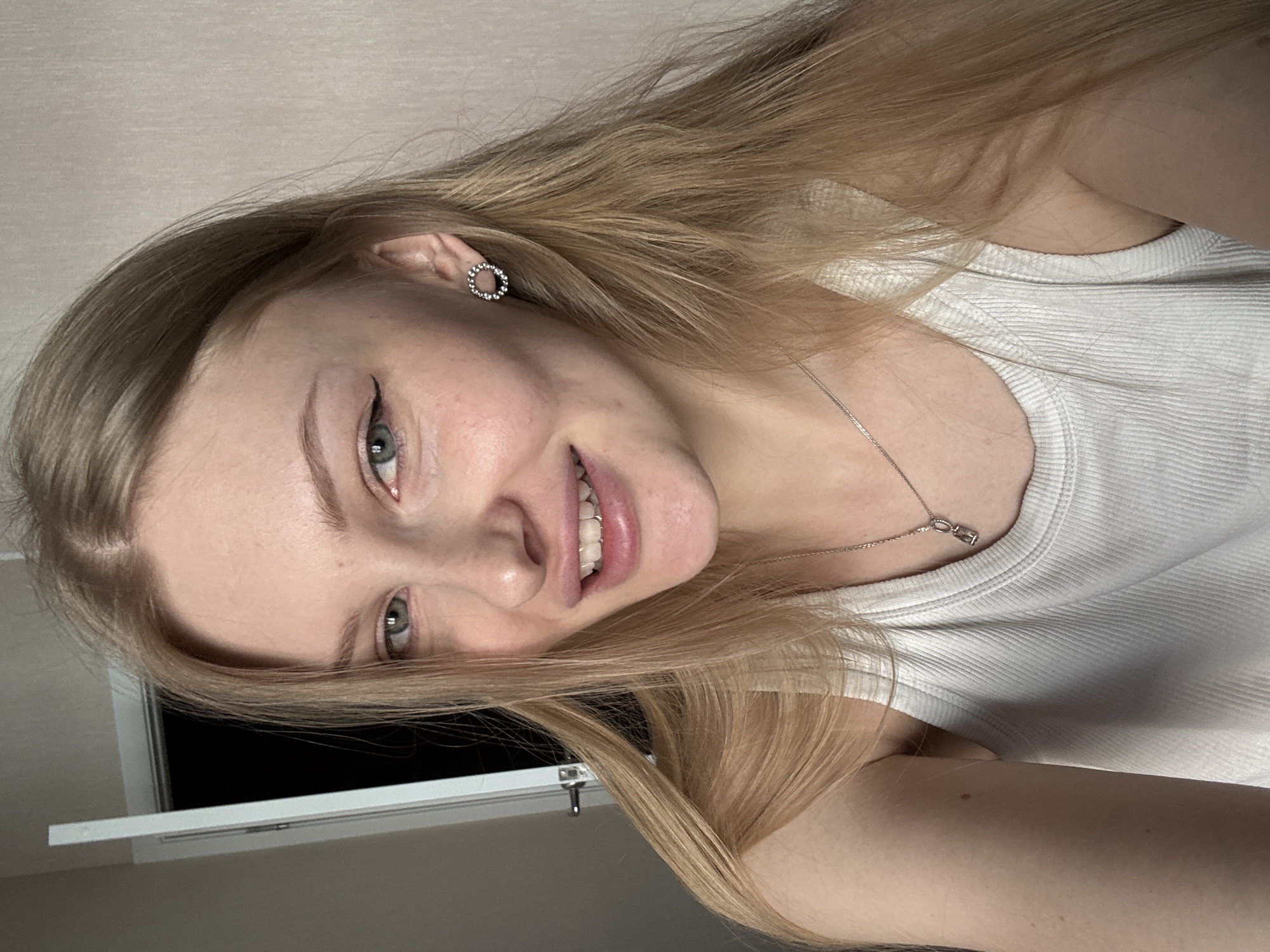 A close-up portrait shows a young woman with long blonde hair and fair skin. She is smiling slightly, revealing her teeth. She wears a white ribbed tank top and a delicate silver necklace with a small pendant. Her eyes are green, and she has eyeliner applied. She has small, circular earrings in her pierced ears. The background is a neutral beige wall, with a glimpse of what appears to be a window frame in the lower left corner. There is no location in Perm, Russia, visible in this image.