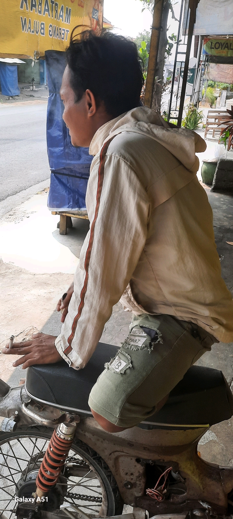 A man with dark hair is seated on a motorcycle in what appears to be an outdoor urban setting, possibly near a roadside stall. He is looking away from the camera, towards the left. He is wearing a light-colored hooded jacket with a reddish-brown stripe down the sleeve and green pants with patches. The man is holding a cigarette in his left hand, with smoke rising from it.
The motorcycle is partially visible, showing the rear wheel, suspension with an orange spring, and part of the seat. The background includes a yellow sign with text that is difficult to read, a blue tarp covering an object, and some foliage. To the right of the man, there are hints of other structures and plants, suggesting an outdoor marketplace or workshop. The ground is a paved surface. The image also displays a watermark in the bottom left corner: "galaxy A51".
The location is identified as Tangkil, Indonesia. The overall impression is of a casual, everyday scene taking place on a street in Indonesia.