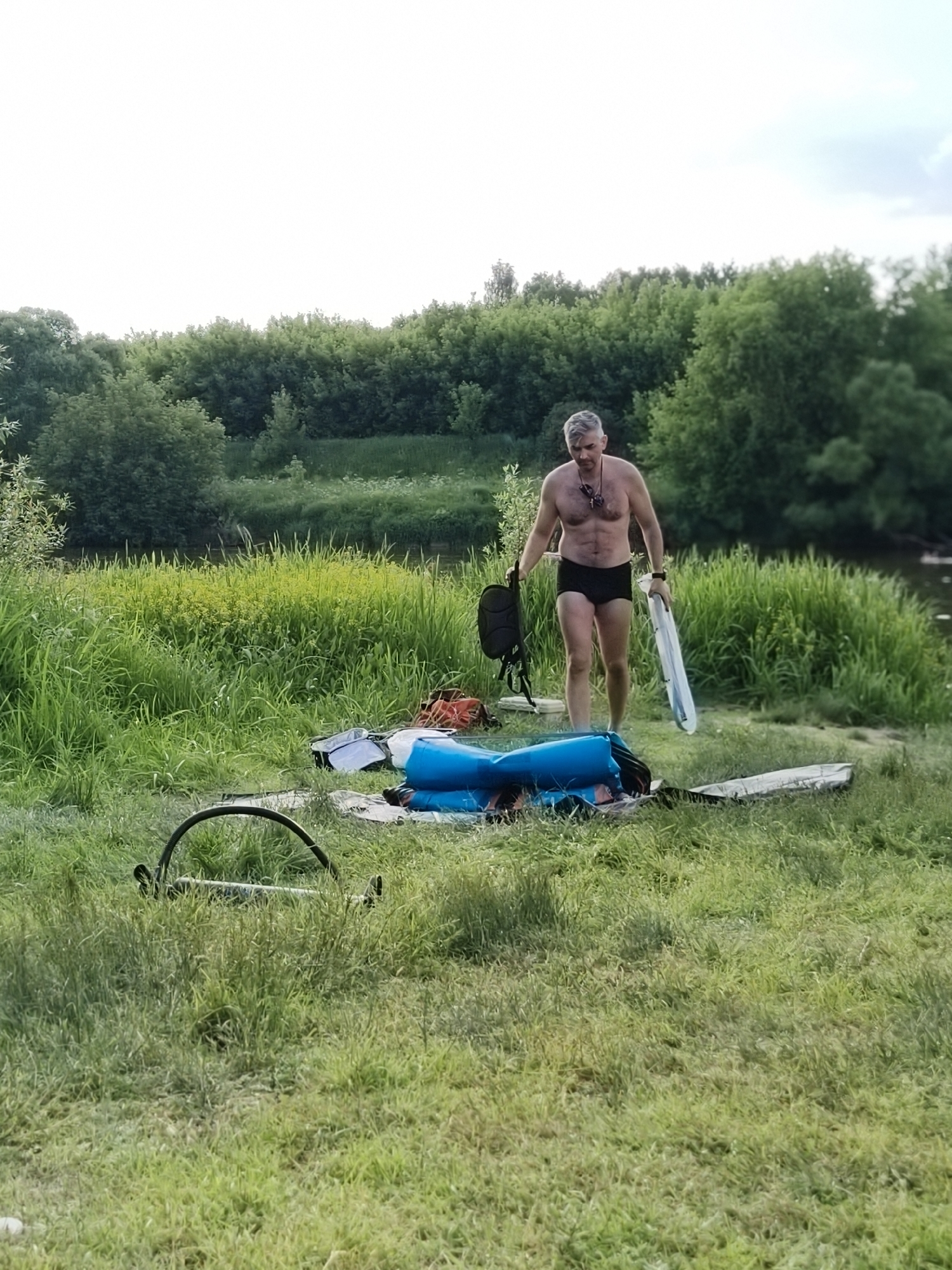 A man in black swim trunks stands in a grassy area near a body of water. He is holding a black backpack in his left hand and a white, flat object in his right. In front of him, on the ground, is a deflated blue inflatable object and other assorted items, including bags and what appears to be a rolled-up tarp or mat. The man is looking downwards. The background consists of lush green trees and foliage. The sky is overcast.