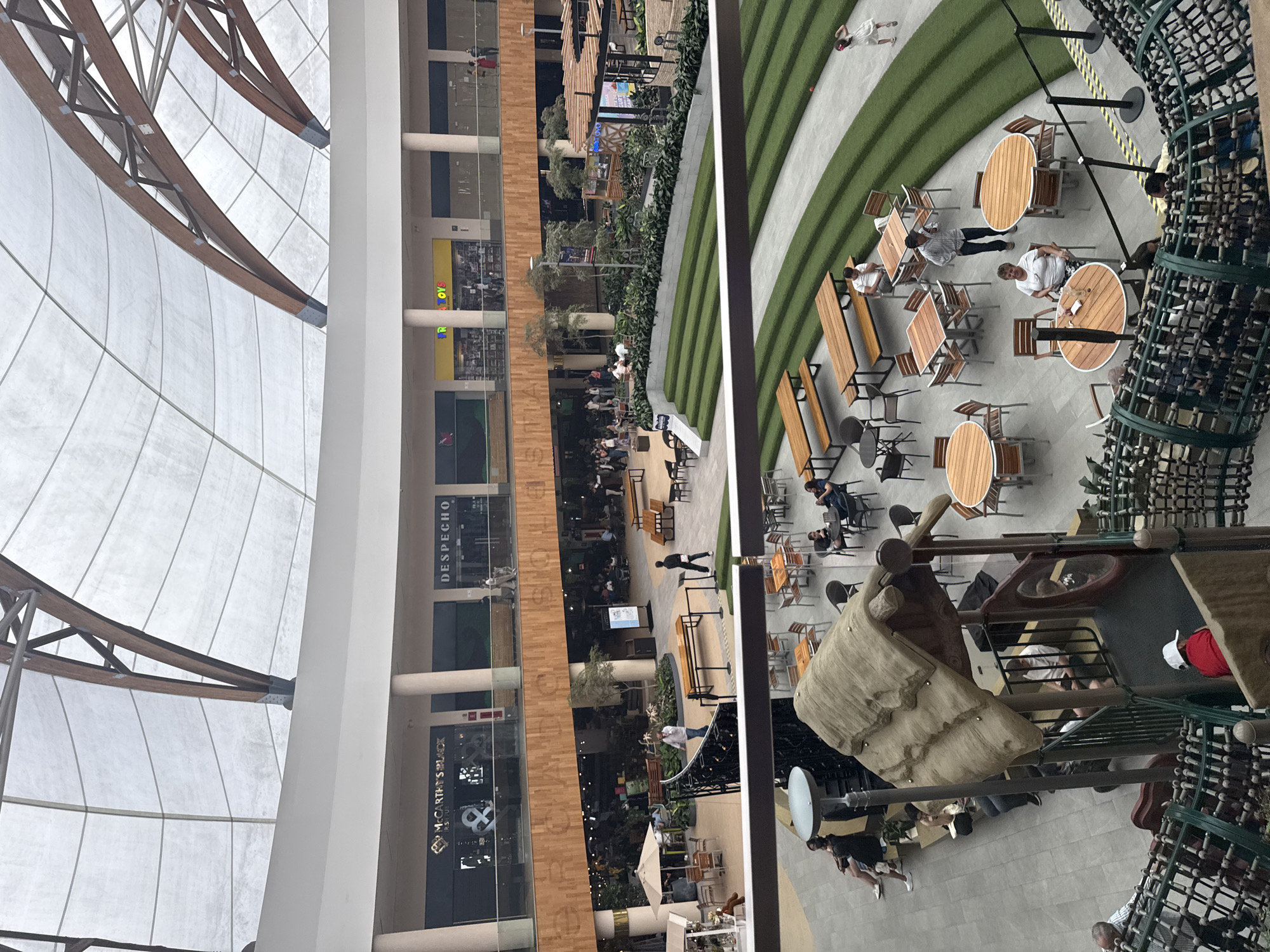The image shows an interior view of a commercial space, likely a shopping mall in Puebla, Mexico. The space features a domed ceiling with wooden beams, which creates a bright and airy atmosphere. The mall has multiple levels with shops and restaurants visible.
On one side of the image, there are storefronts with businesses such as "Despecho" and "McCarthy's Black." People can be seen walking and sitting at tables in the central area.
The other side of the image has outdoor seating areas with tables and chairs, as well as a children's playground with climbing structures. Artificial grass adds a touch of greenery to the space. Several people are seated at the tables, seemingly relaxing or dining, while others are playing in the playground area.
The image captures a bustling and lively atmosphere in a modern and well-designed commercial space in Puebla, Mexico.