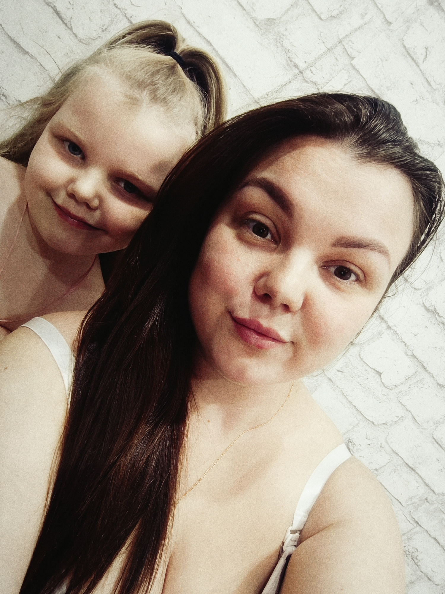 A selfie of a woman with long brown hair and a young girl with blonde hair pulled back in a ponytail. The woman is in the foreground, smiling slightly. The young girl is behind her, also smiling. The background is a light-colored brick wall. It's not possible to confirm the specific location within Chaikovsky, Russia based on the image alone.