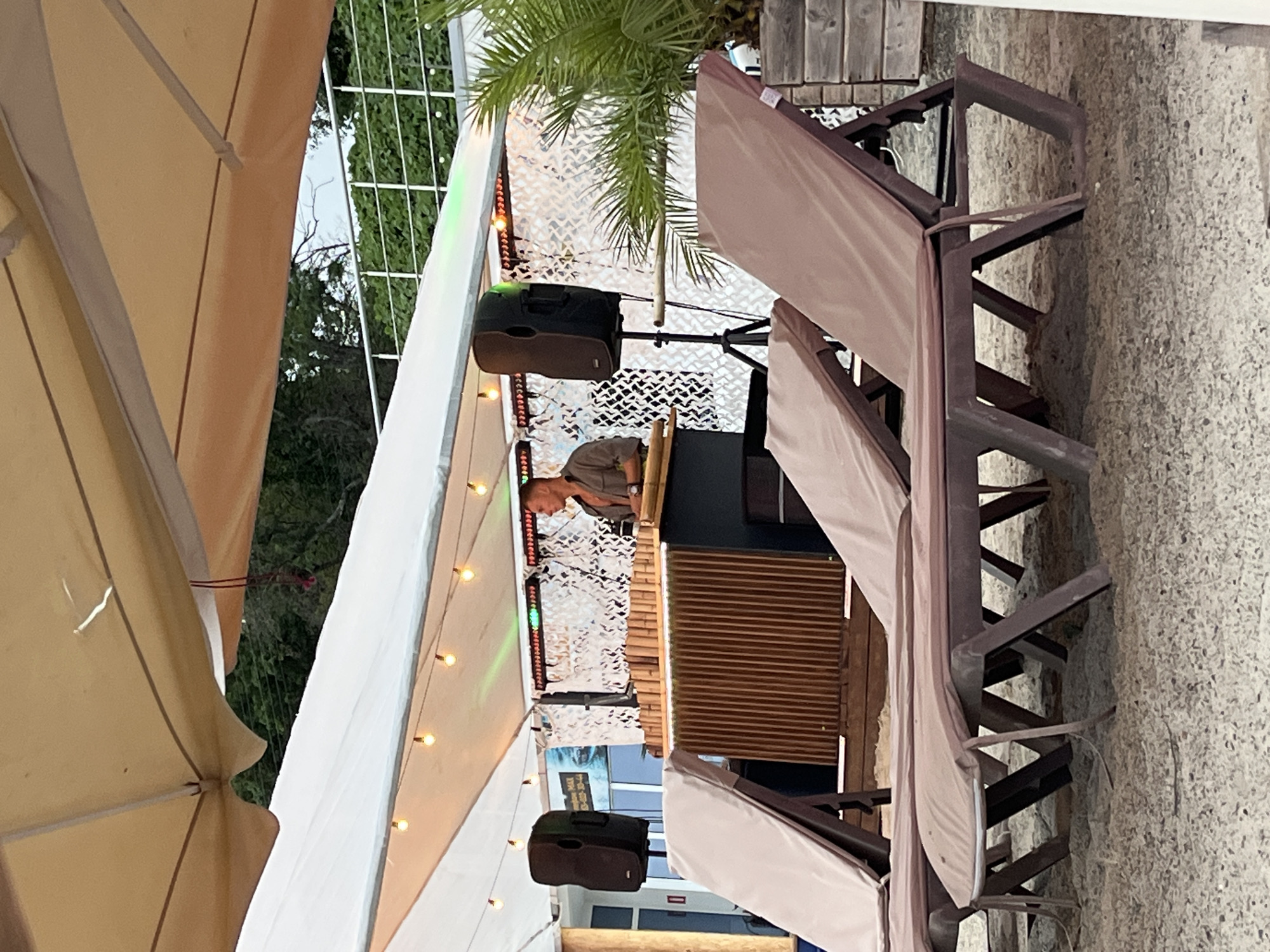The image depicts an outdoor scene at a beachside venue, likely a bar or club, under a large canopy. In the background, a male DJ is positioned behind a bamboo-clad bar, actively engaged with equipment. Two large speakers are visible, one on either side of the DJ booth. The area is decorated with string lights and a white camouflage-patterned backdrop. In the foreground, several empty sun loungers are arranged on the sand, suggesting a leisurely environment. The overall ambiance suggests a daytime event with entertainment. The location is identified as Sochi, Russia.
