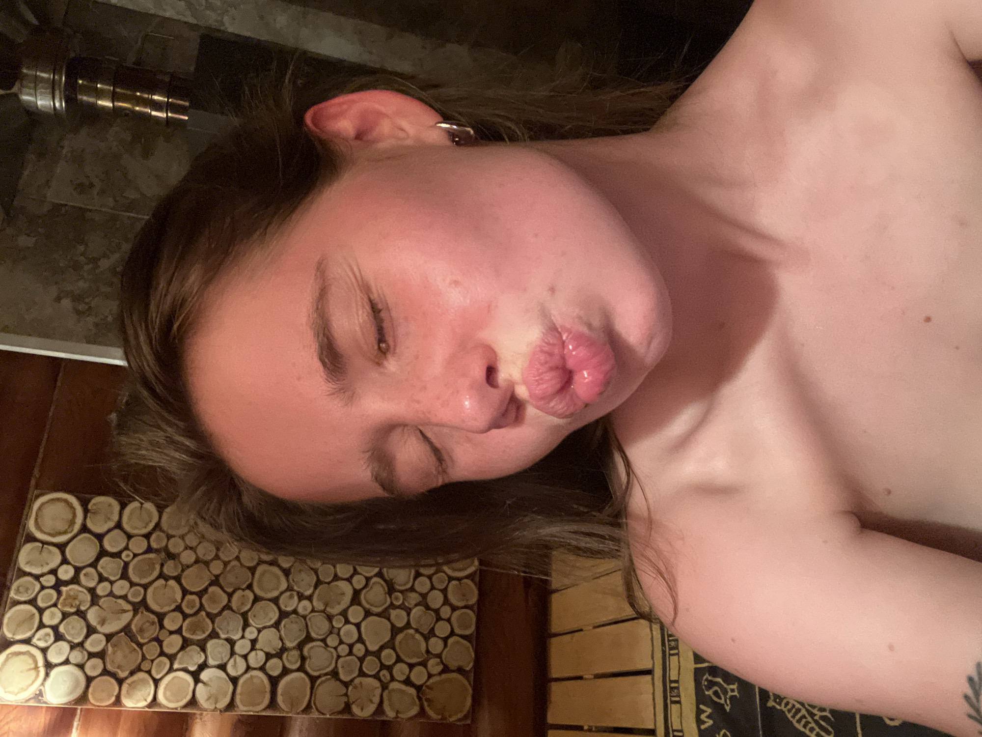 A close-up, horizontally-oriented image depicts a fair-skinned person with their eyes closed, puckered lips, and freckles on their face. They have brown hair and are wearing a hoop earring. The person is lying on a surface or leaning against a wall covered with wood and a shower head. The location is not discernible.