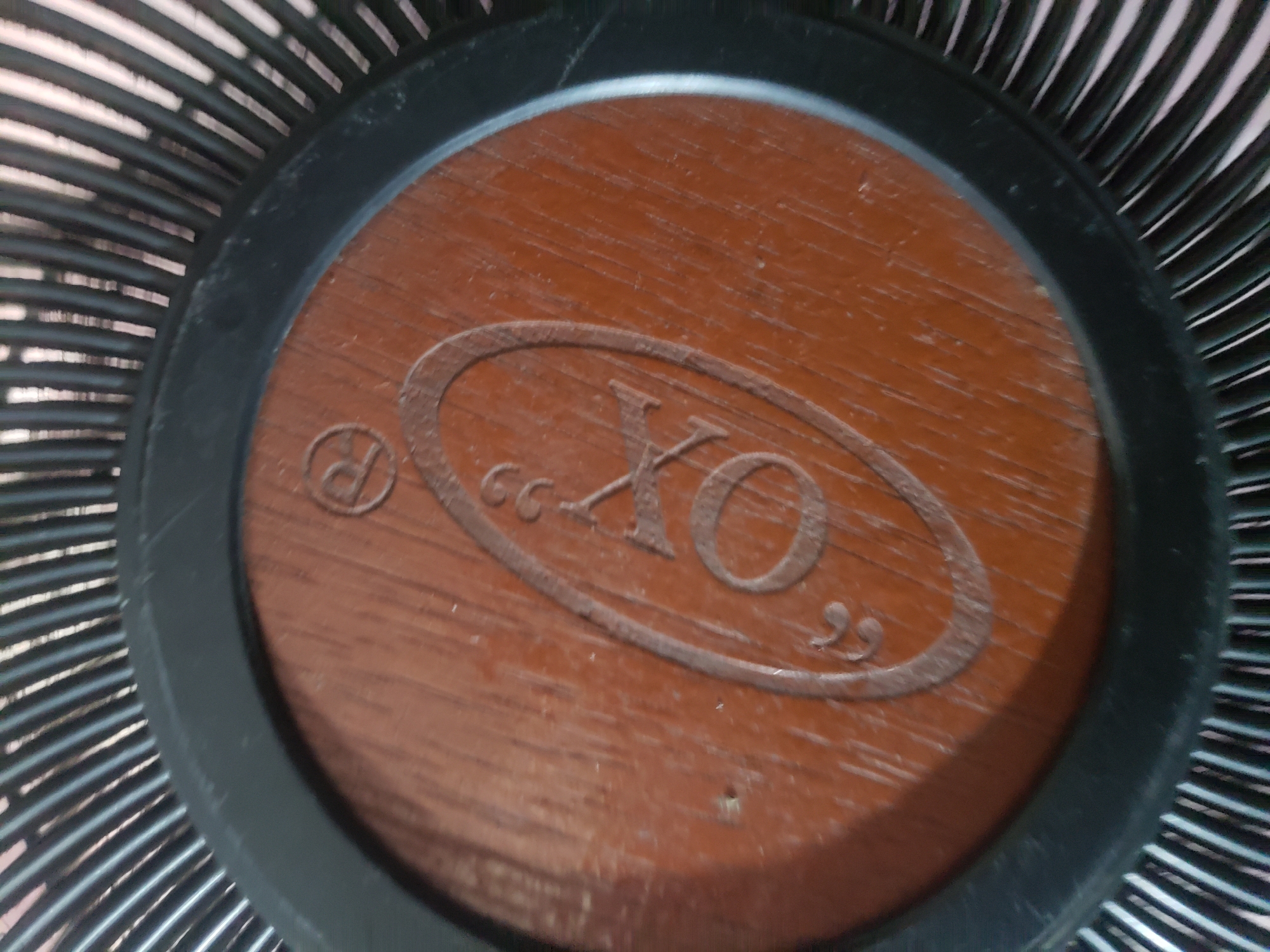 The image shows a close-up of a fan guard with a wooden-like circular emblem in the center. The emblem has decorative flourishes on the left and right sides and appears to have the letters "OX" embossed in the center, possibly with a registered trademark symbol to the right of the "X". The fan guard itself is black and made of thin, parallel metal wires. The background is out of focus, showing more of the fan guard's structure. The image is taken from a slightly elevated angle, looking down at the fan emblem. There are no people or identifiable landmarks present.