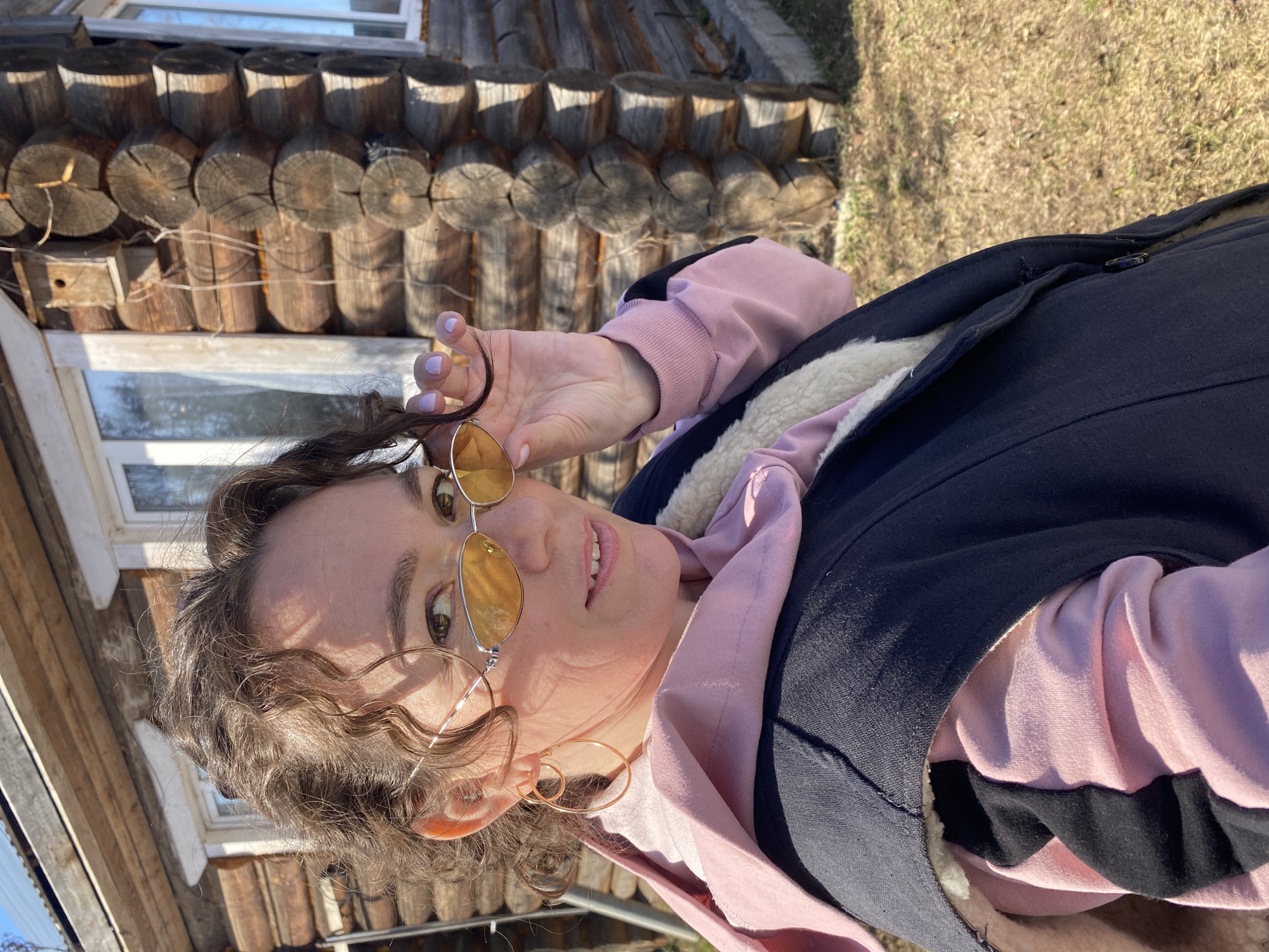 A fair-skinned woman with curly brown hair is posing outdoors, holding the frames of her sunglasses in her right hand. She is wearing gold hoop earrings, a pink hoodie, and a dark vest with a fur lining. In the background, there is a log cabin with visible windows and a birdhouse. The ground is covered with dry grass and dirt. The location is likely near a rural or wooded area in Ольховка, Russia.