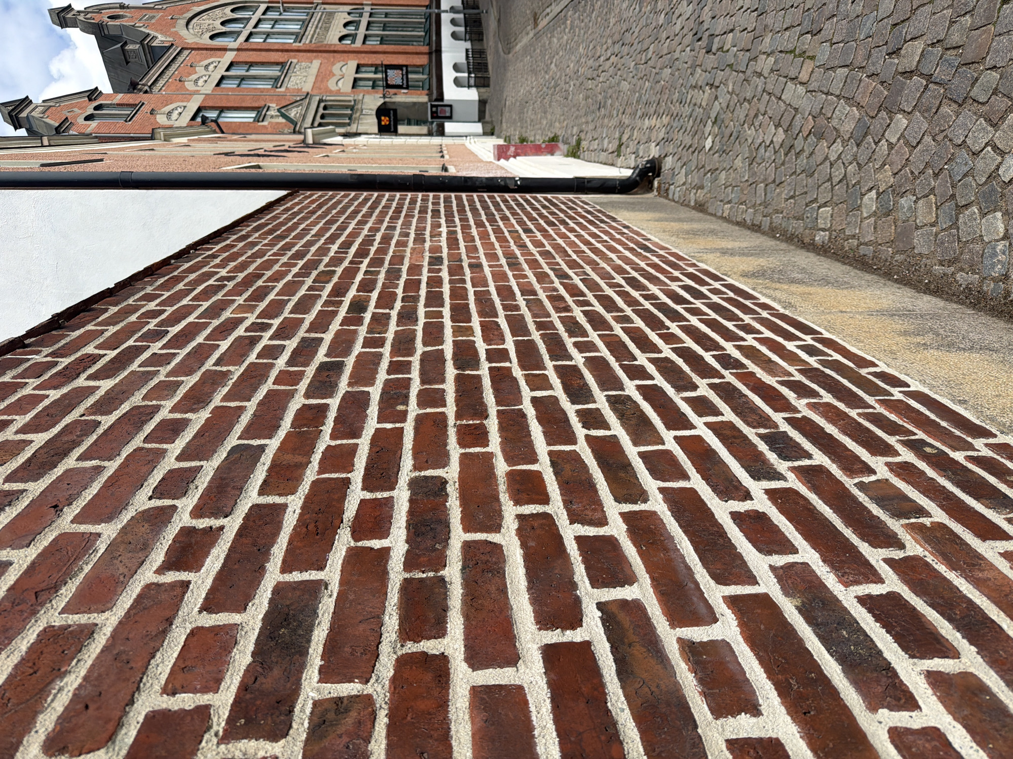The image shows a cobblestone street in the city of Lund, Sweden, with a prominent red brick wall on the left and historic buildings on the right. The brick wall, made of dark red bricks with light grey mortar, fills a significant portion of the foreground and extends into the background. The street is paved with irregular-shaped grey cobblestones.
In the background, to the right of the brick wall, are several buildings with distinctive architecture. The closest building is an ornate, multi-story structure made of red brick with white trim and dark arched windows. Above it, another building with a dark, sloped roof and pointed architectural features is visible. Several signs are mounted on the buildings, including a dark sign with a crest and another smaller, darker sign. A black downspout runs vertically along the side of the brick wall. The sky is partly cloudy.
No people are visible in the image. The scene is a street-level view, likely taken during the daytime given the lighting. The perspective is from low down, looking diagonally along the street and wall.