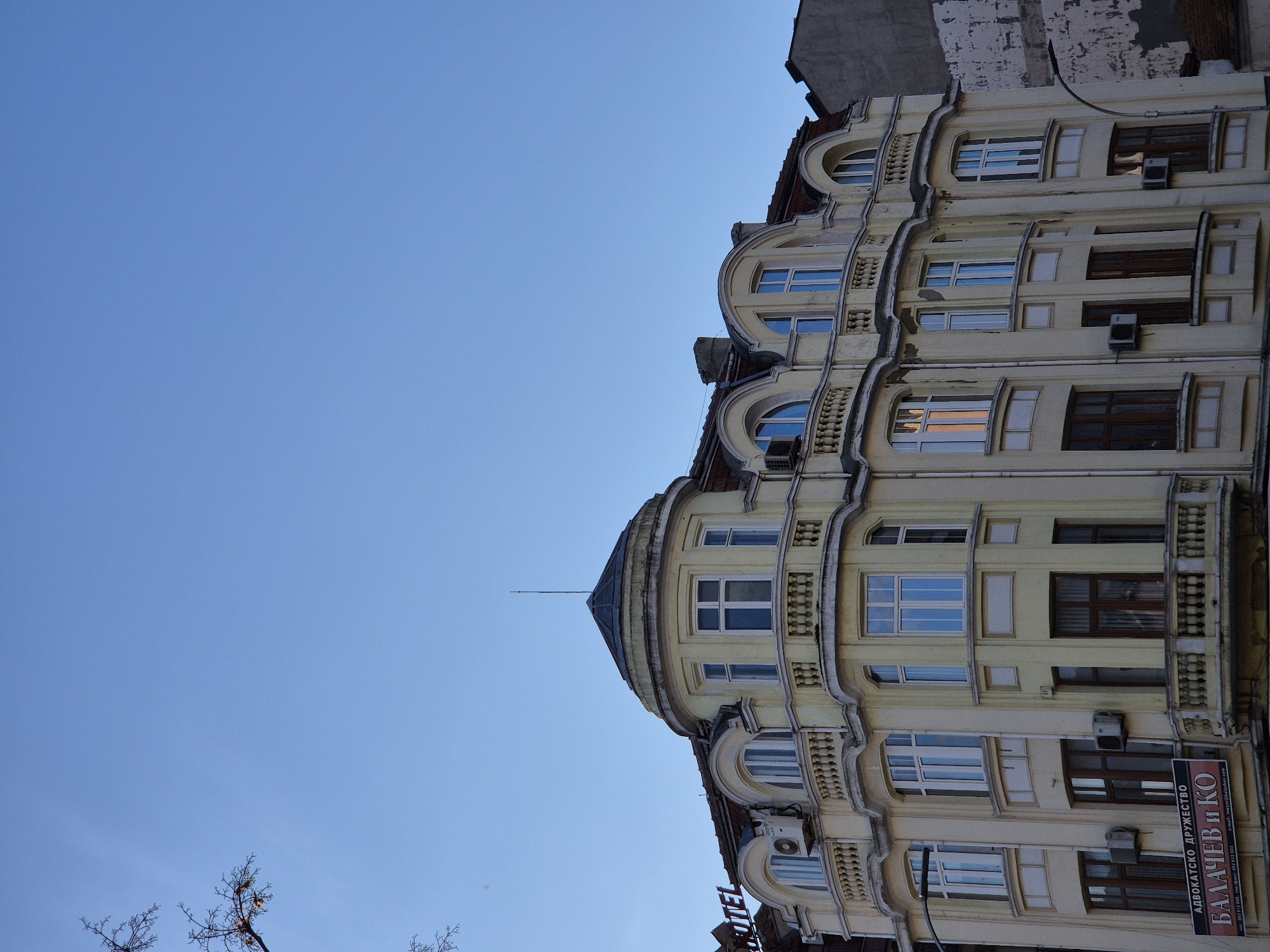 The image presents a low-angle view of a multi-story building in Varna, Bulgaria. The building features a cream-colored facade with ornate, classical-style window detailing and decorative moldings. The architecture includes arched windows on the upper floors and a rounded turret at the corner, topped with a pointed roof and lightning rod. Windows are a mix of closed and partially open. A small sign with the text "HOTEL" is barely visible near the bottom left. Below the building the text "АДВОКАТСКО ДРУЖЕСТВО БАЛАЧЕВ И КО" is also visible. The sky above is clear and blue, while bare tree branches extend into the frame from the left. A fragment of an adjacent building is visible at the top of the frame.