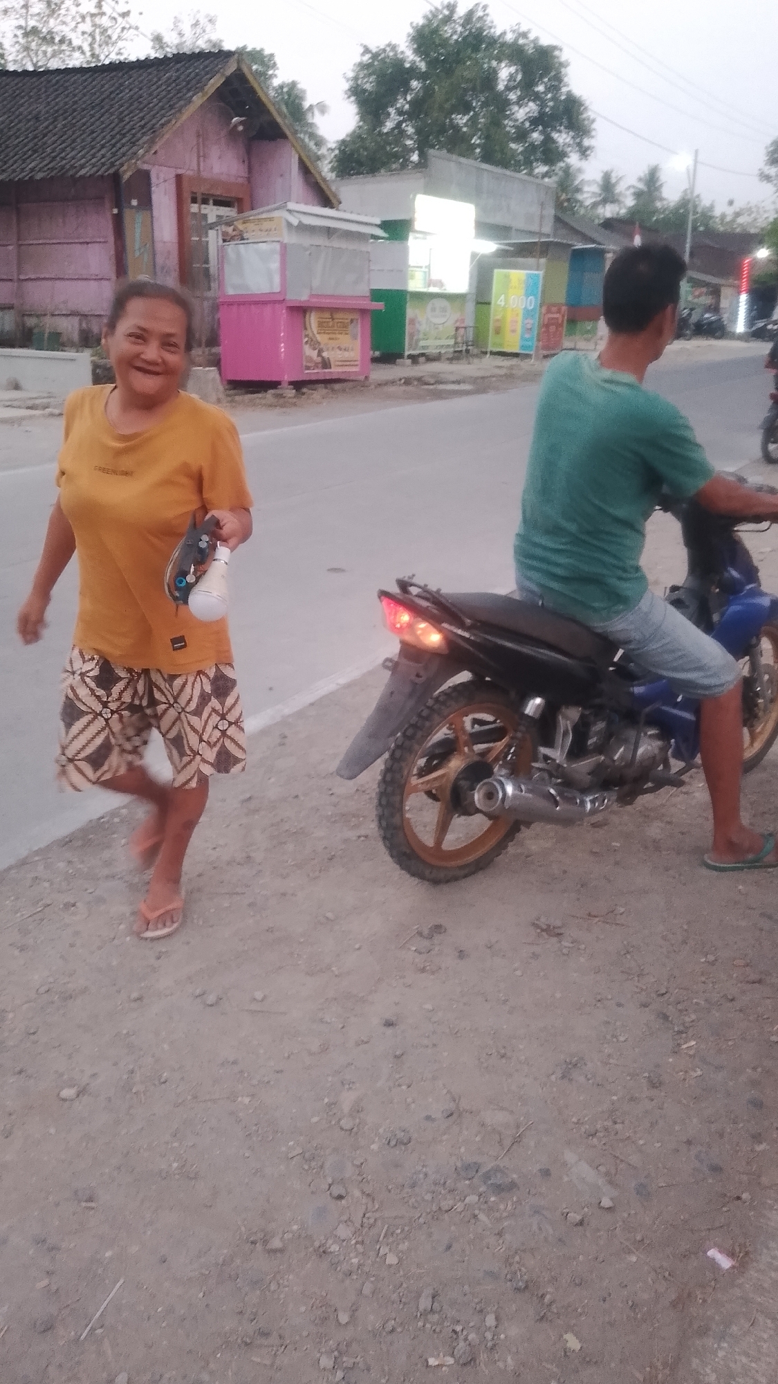 A woman in an orange t-shirt and patterned shorts smiles at the camera, holding a lightbulb and a controller in her left hand. She is standing on a dirt road next to a motorcycle parked on the side of the road. A person wearing a green t-shirt and denim shorts is sitting on the motorcycle. In the background, there are several small shops and buildings. The scene appears to be in Karanglangu, Indonesia, as indicated by the visual elements and context.