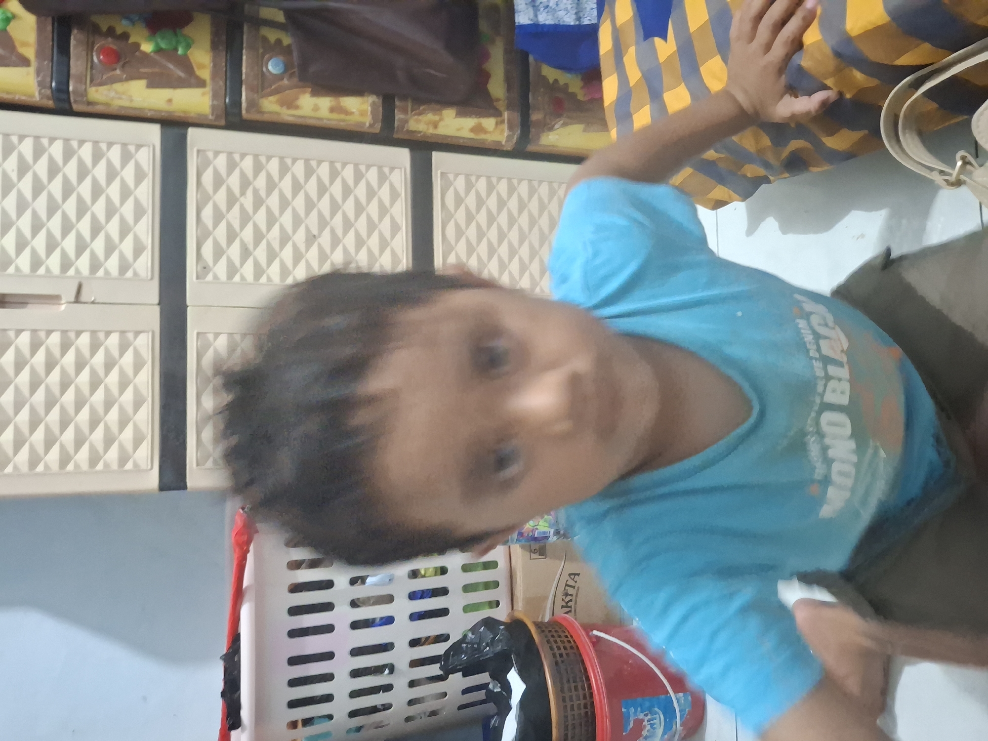 A young boy is kneeling on a tiled floor in front of a yellow and blue striped mat. He is wearing a blue t-shirt with white text that reads "MONO BLAC". He has dark, spiky hair and is looking forward with a blurry expression. Behind him, there is a white laundry basket with some black plastic bags in it, and a red bucket. Further back, there is a beige and brown plastic cabinet with multiple drawers. The wall behind the cabinet is light blue. The scene appears to be indoors, likely in a residential setting in Biak, Indonesia.
