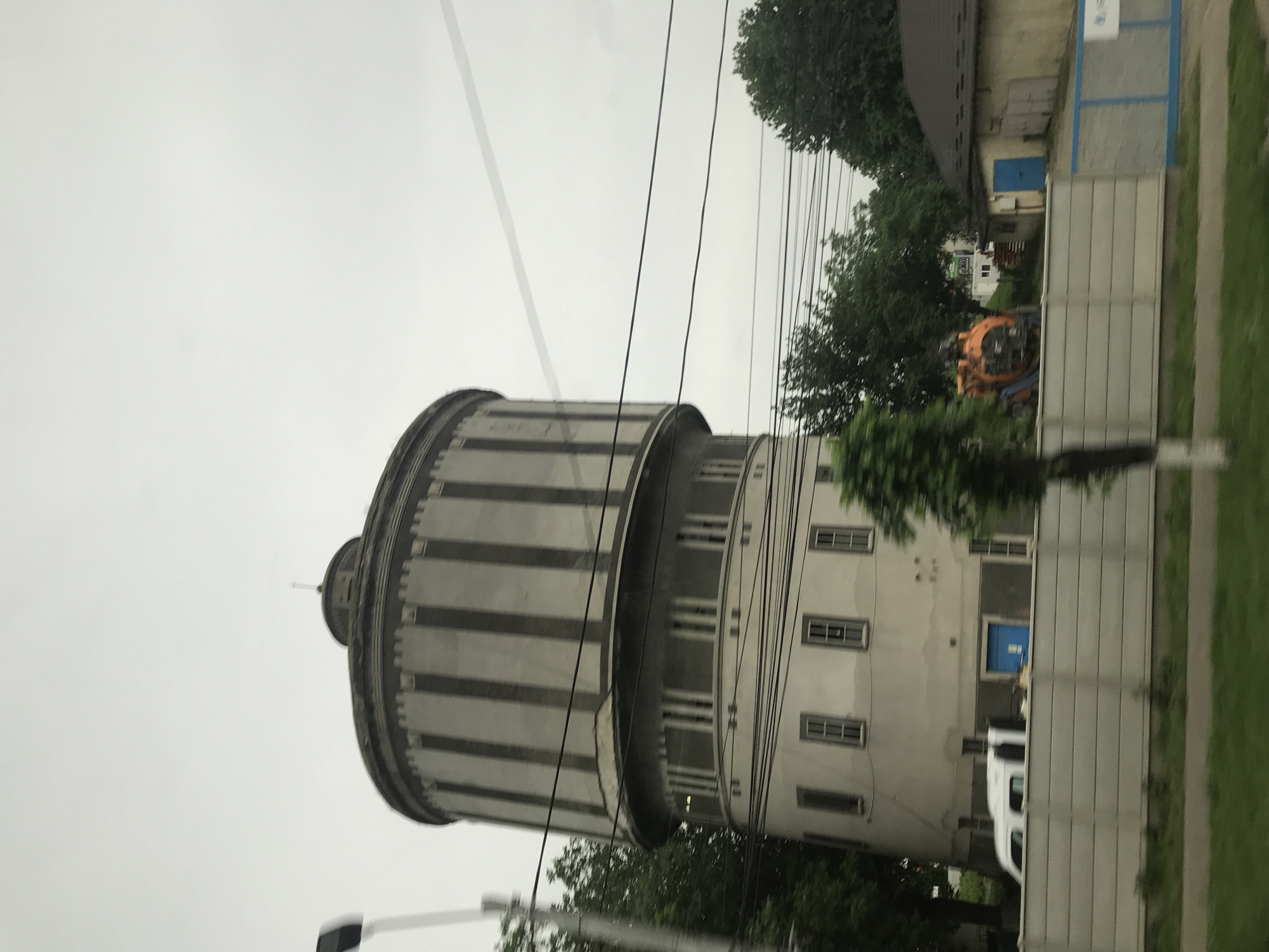 This image captures a multi-tiered cylindrical building, identified as a historic water tower, situated in the city of Roman, Romania. The upper section of the tower features a dark grey cylindrical tank adorned with prominent, regularly spaced vertical dark grey pilasters or decorative bands, capped by a shallow domed roof and a small, slender finial at its apex. Beneath this, a narrower, light grey cylindrical level displays a series of evenly spaced tall, narrow rectangular windows. The widest base section of the tower is light beige, presenting several grey-framed rectangular windows and a recessed blue door at its base on the left side.
In the immediate foreground, a strip of green grass runs along the bottom, bordered by a light grey concrete panel fence that stretches across the scene. A utility pole is partially visible on the far left. Behind the fence, a white panel van is parked towards the left, while further right, a large orange piece of heavy machinery, possibly an excavator or industrial pump, stands next to a smaller, single-story building with a brown-tiled roof and a bright blue door. Abundant green trees are visible around and behind the structures. Numerous black power lines crisscross the middle ground against an overcast, light grey sky. There are no individuals present, and no actions or interactions are depicted.