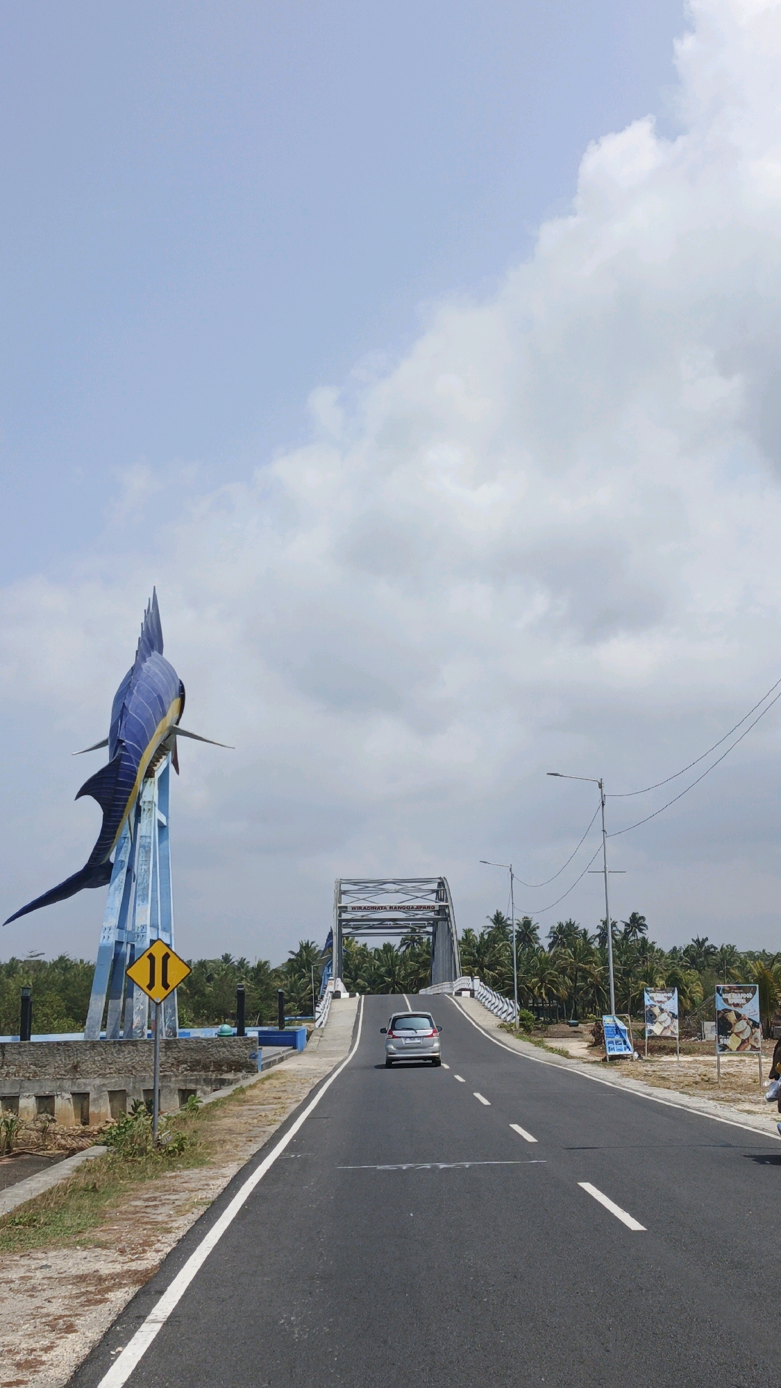 The image depicts the Grand Marlin Statue, a prominent landmark in Pangandaran, Indonesia. The statue, a large blue marlin with yellow accents, is positioned on the left side of the frame, supported by a blue structure. In the foreground, a paved road stretches towards a bridge, with a silver car driving across it. The bridge is an arch bridge with the text "WK. ADINATA RANGGA SIPANG" visible on its structure. The surrounding environment is lush with palm trees, and the sky is filled with scattered clouds. There are also several billboards on the right side of the road, some featuring images of food. A yellow traffic sign with a diagram of two converging lines is also visible near the statue, indicating a road narrowing.